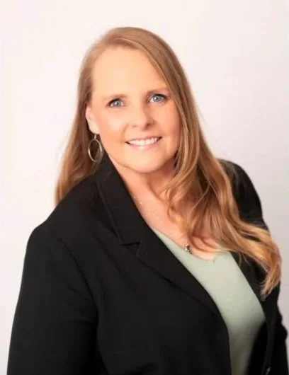 Portrait of a woman with long, wavy blonde hair, wearing a black blazer and light green top, smiling at the camera against a plain background.