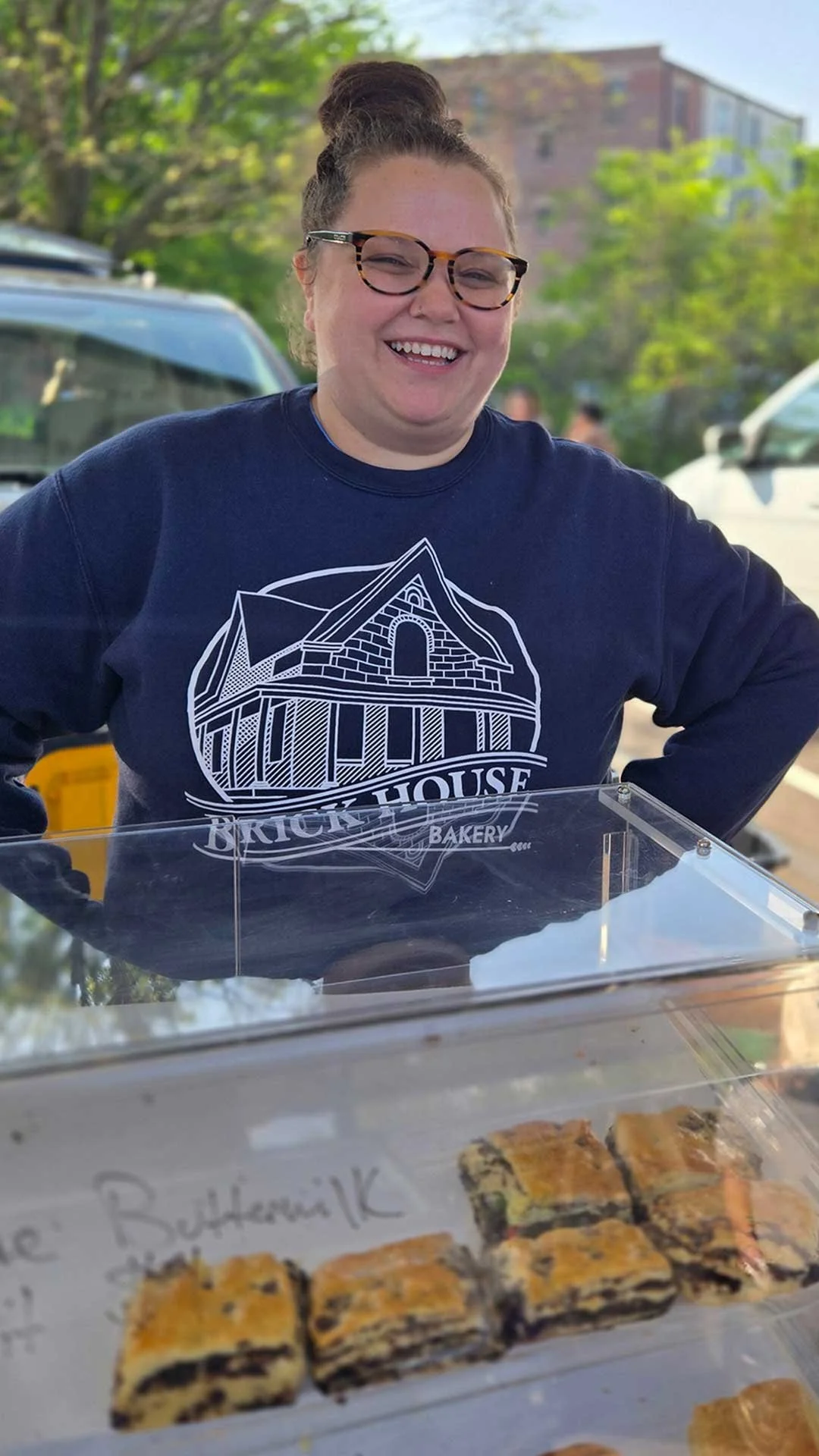 Smiling woman with glasses wearing a navy blue sweatshirt that says "Brick House Bakery." She is standing outdoors with trees and cars in the background. In front of her, there is a clear display case with baked goods.