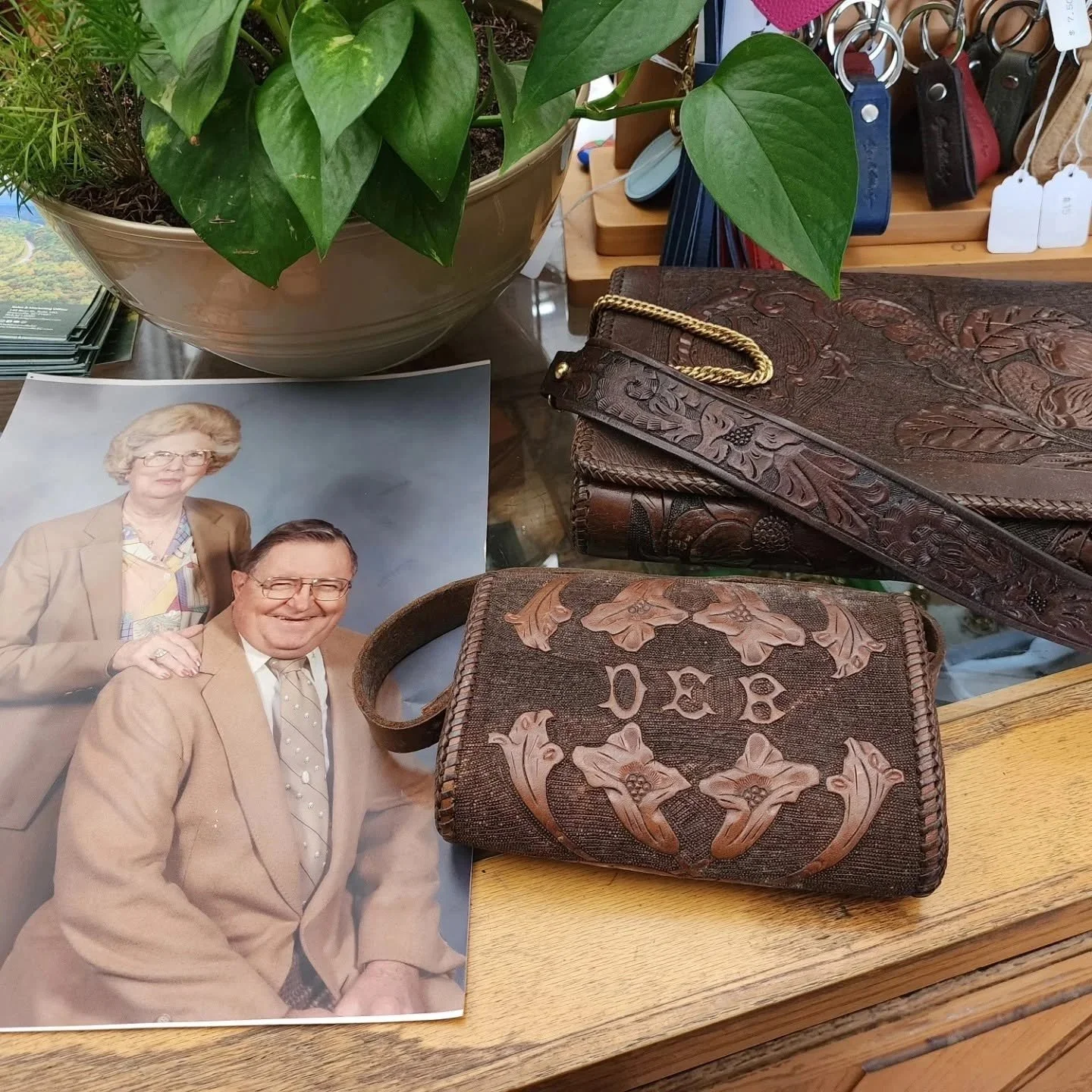 Photograph of two elderly people, a woman and a man, dressed in beige suits, smiling. Items on a wooden surface include two vintage leather wallets with embossed floral patterns, a purse with a leather strap, a large potted green leafy plant, and a display of keys and tags in the background.