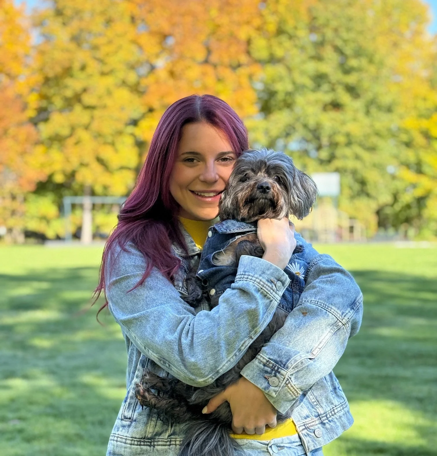 A young woman with purple hair holding a small gray dog in her arms, standing outdoors in a park with autumn-colored trees in the background.