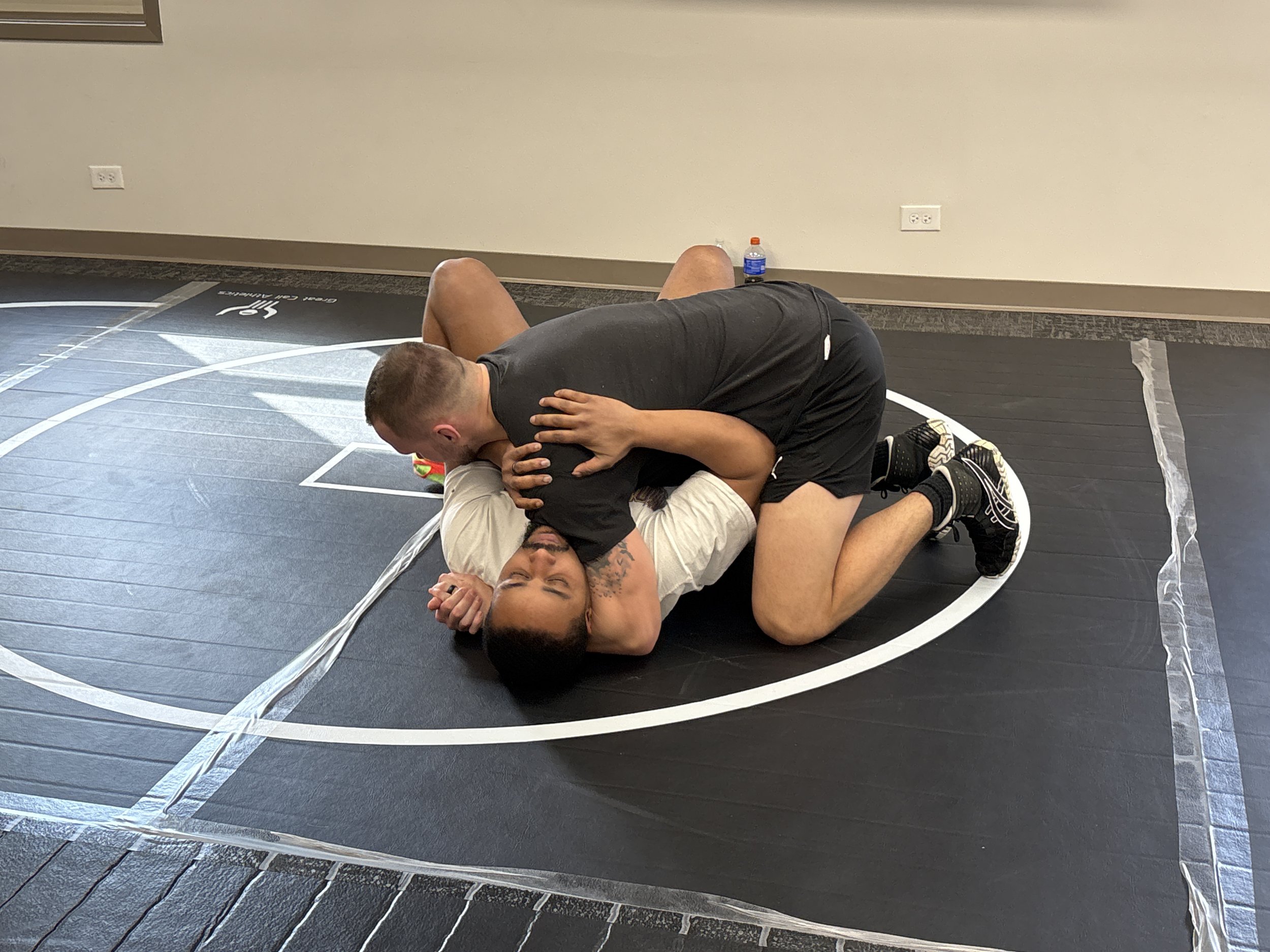 Two men practicing Brazilian jiu-jitsu on a black wrestling mat inside a gym, with one man on top of the other in a grappling position.