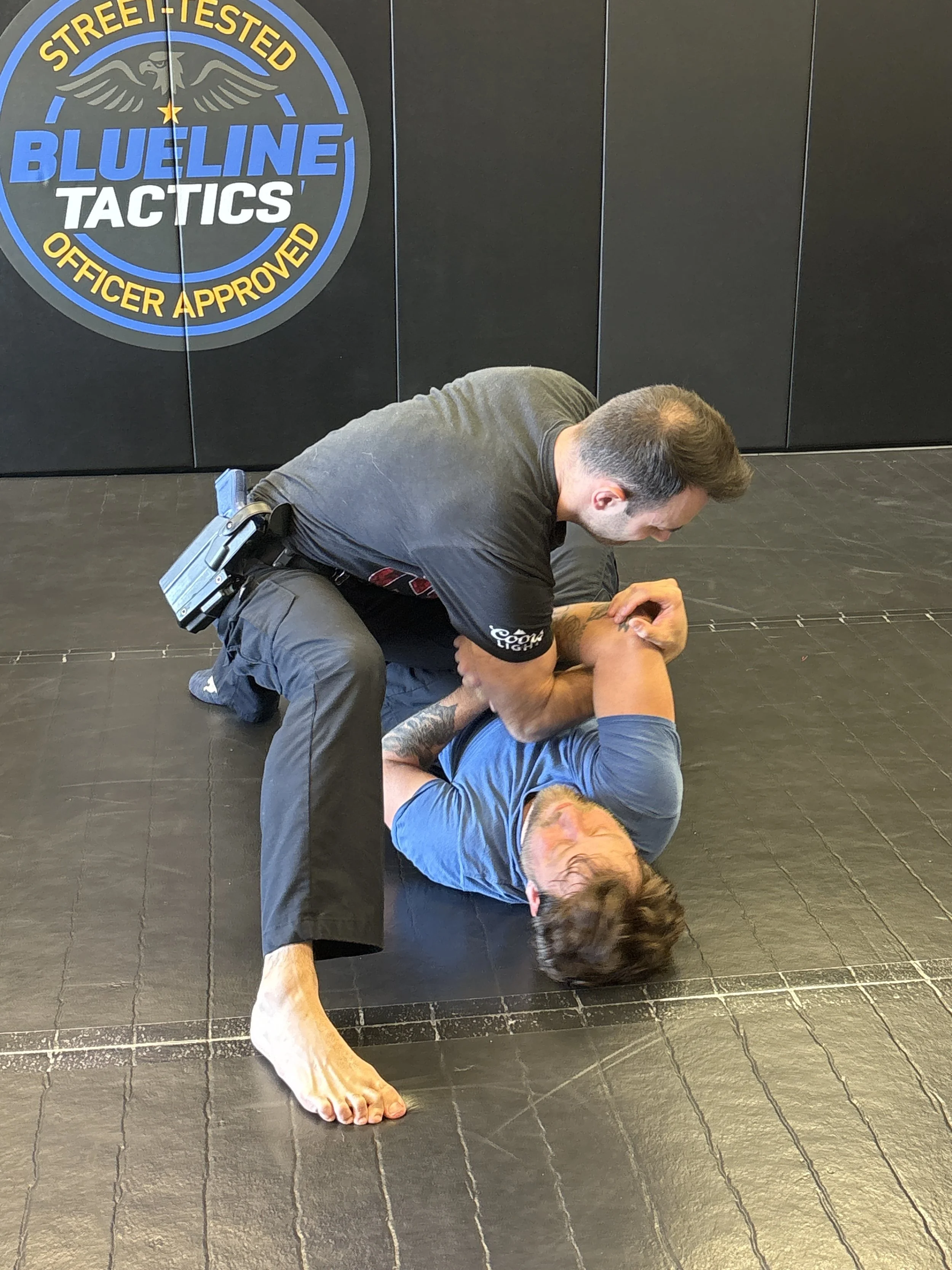 A woman in a gray shirt practicing Brazilian jiu-jitsu on a man in a blue shirt on a black mat. An officer in a black uniform is pinning the woman to the ground.