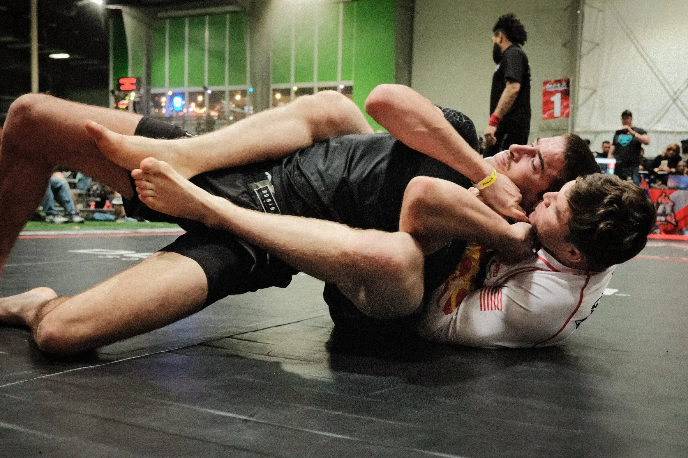 Two male competitors engaged in a grappling match on a wrestling mat at an indoor sports facility, with one on top of the other holding each other closely, in the background other spectators and officials watch, and a person with a beard and a ponytail stands observing the match.