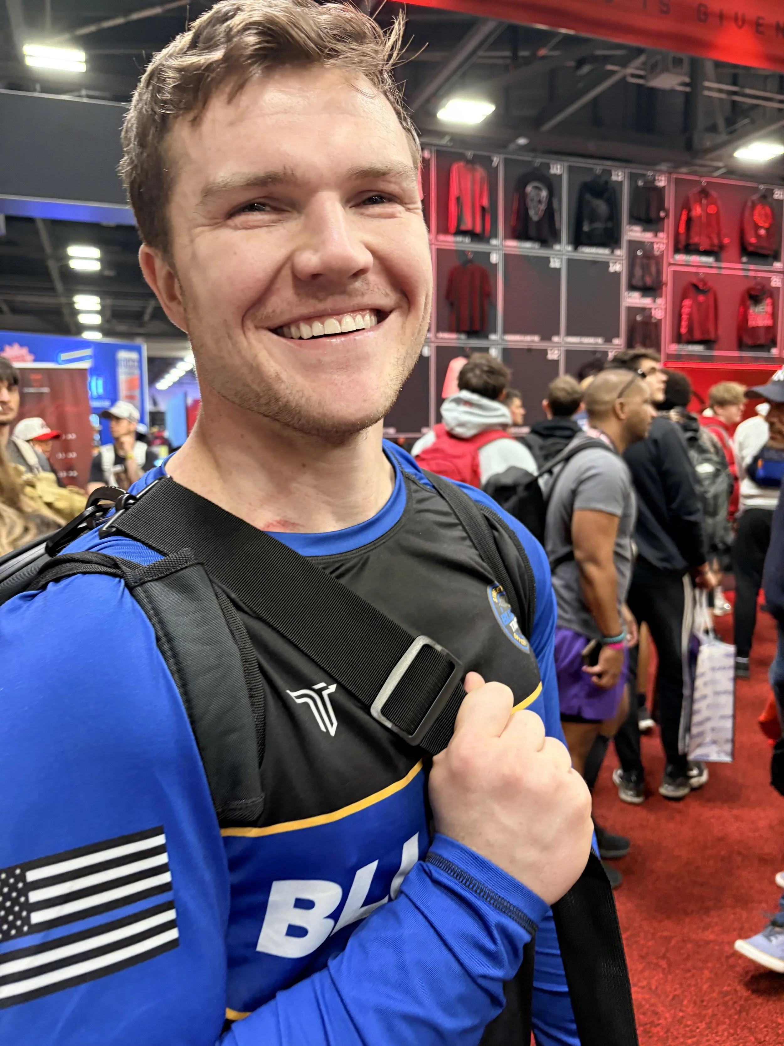 A young man with short brown hair smiling at the camera, wearing a blue sports jersey and a black backpack, standing in a crowded indoor area with people in line and a display of clothing in the background.