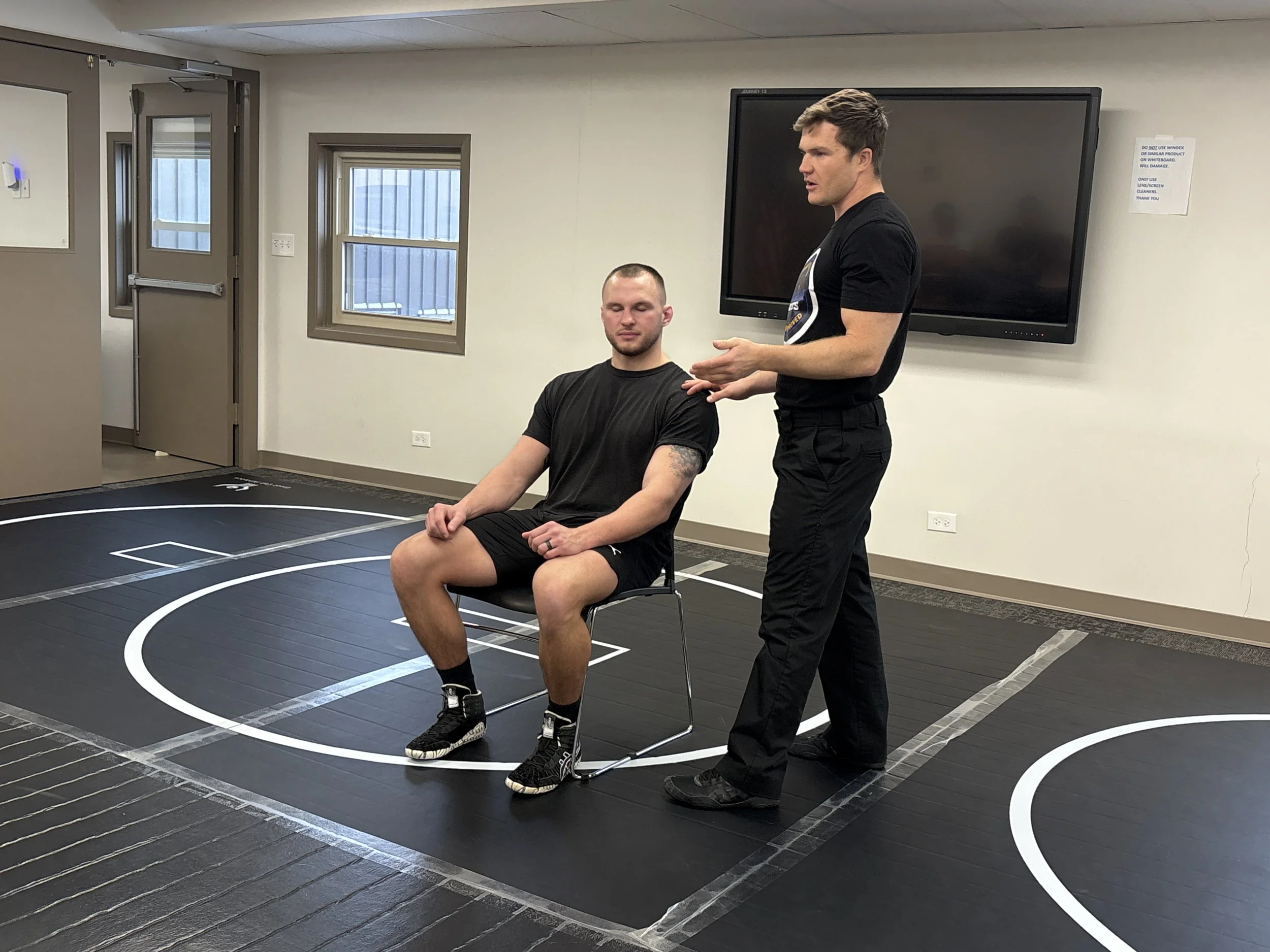 Two men in a room with black flooring marked with white lines. One man is sitting with his eyes closed, wearing a black shirt and shorts, while the other man is standing next to him, talking or explaining something. There is a large black screen mounted on the wall behind them, and two windows with blinds. The room appears to be a gym or training space.