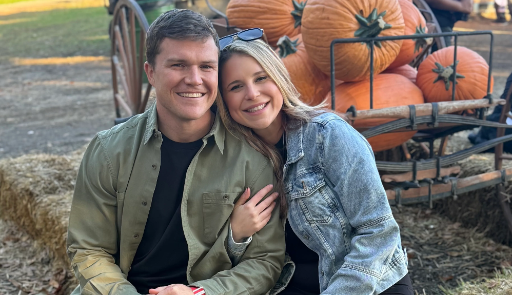 A smiling young couple sitting outdoors near a wagon filled with large pumpkins, with hay bales on the ground, indicating a fall or pumpkin patch setting.