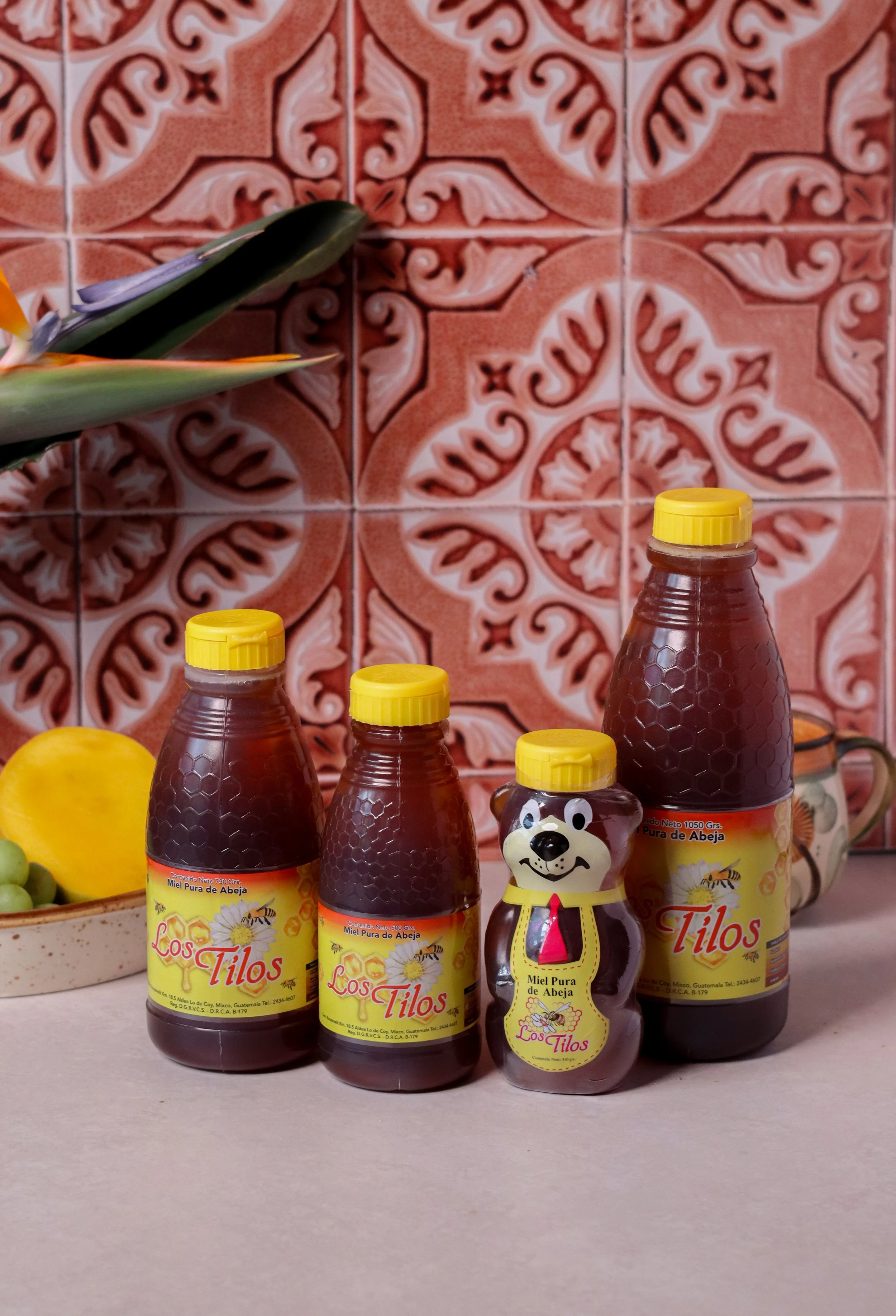 Botellas de miel pura de abeja con etiquetas amarillas, una pequeña figura de perro con una vestimenta amarilla y una abeja, y algunos frutos alrededor, en una encimera con fondo de azulejos decorados rojos y blancos con motivos florales.