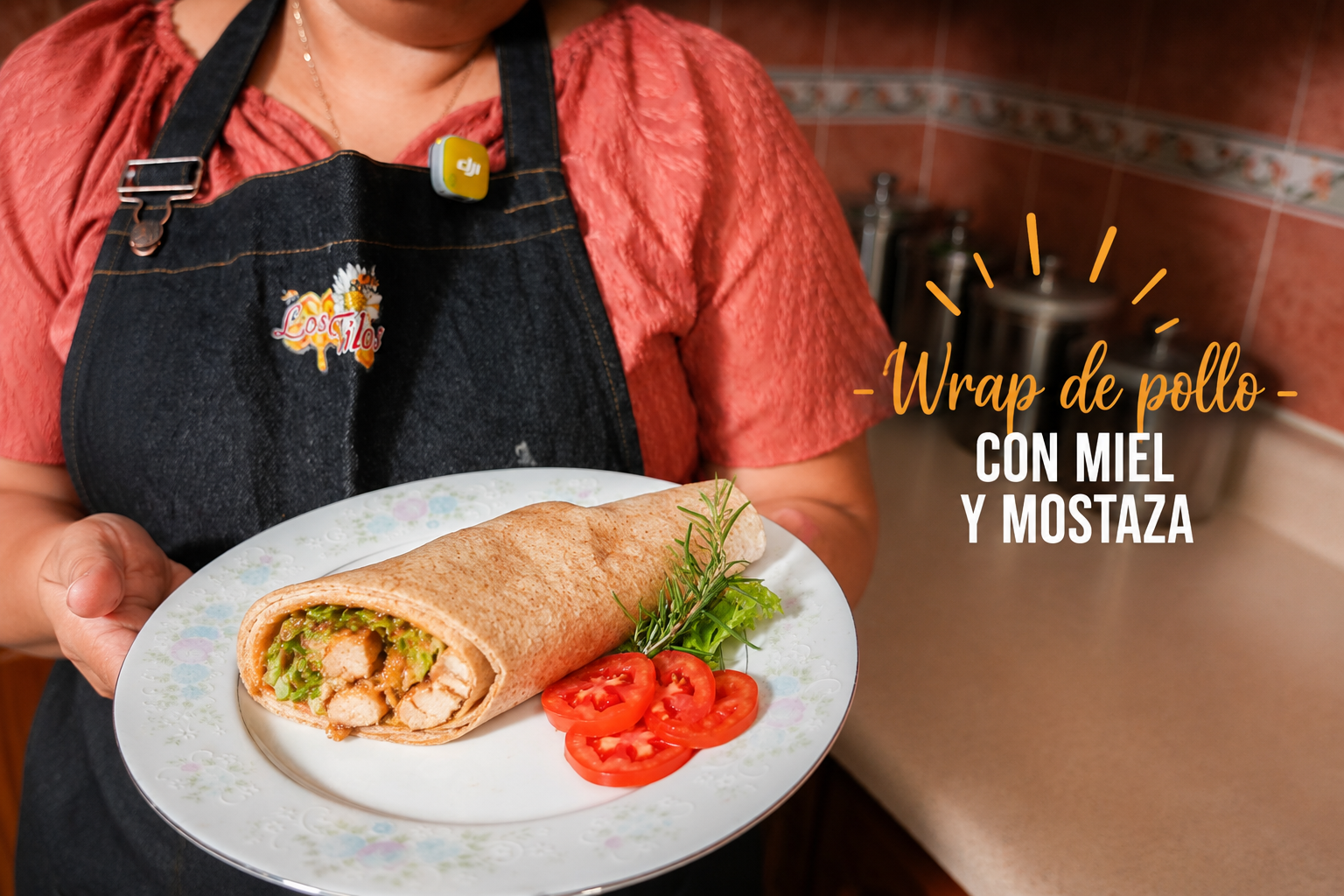 Persona sosteniendo un plato con un wrap de pollo con miel y mostaza, acompañado de tomate y hierbas en un entorno de cocina.