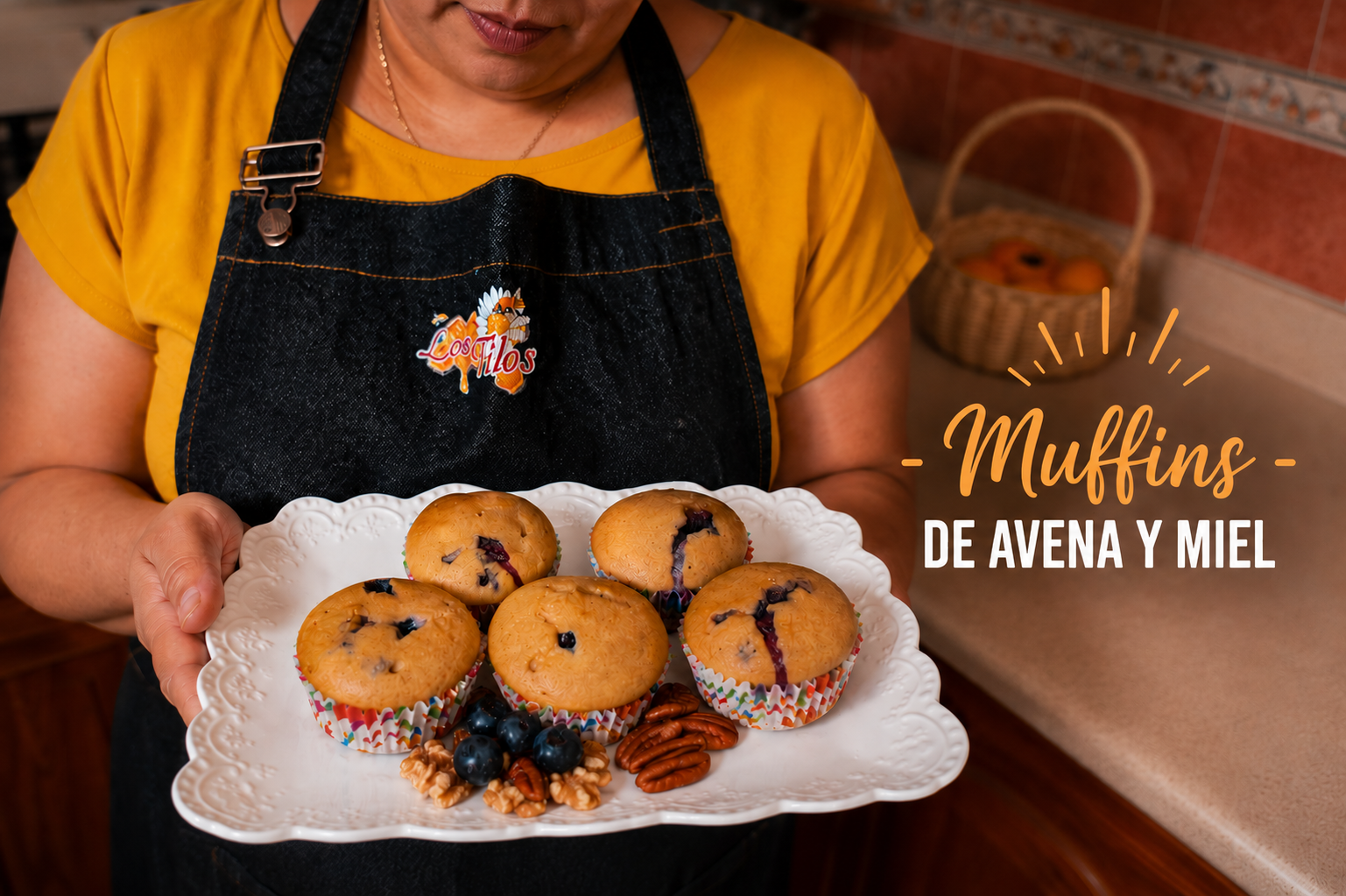 Mujer sosteniendo un plato blanco con cinco muffins de avena y miel decorados con arándanos y nueces, en una cocina. En la esquina derecha, texto que dice 'Muffins de avena y miel'.