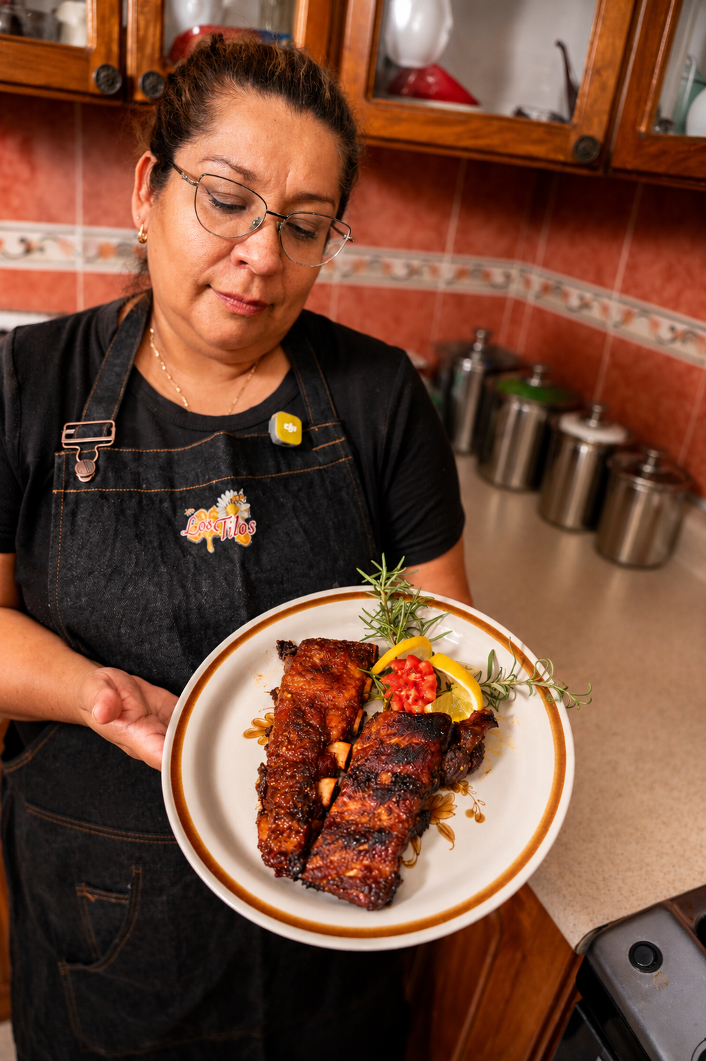 Mujer sosteniendo un plato con costillas a la barbacoa decoradas con ramitas de romero, rodajas de limón, tomate y pimientos en un fondo de cocina con gabinetes de madera y frascos de almacenamiento.