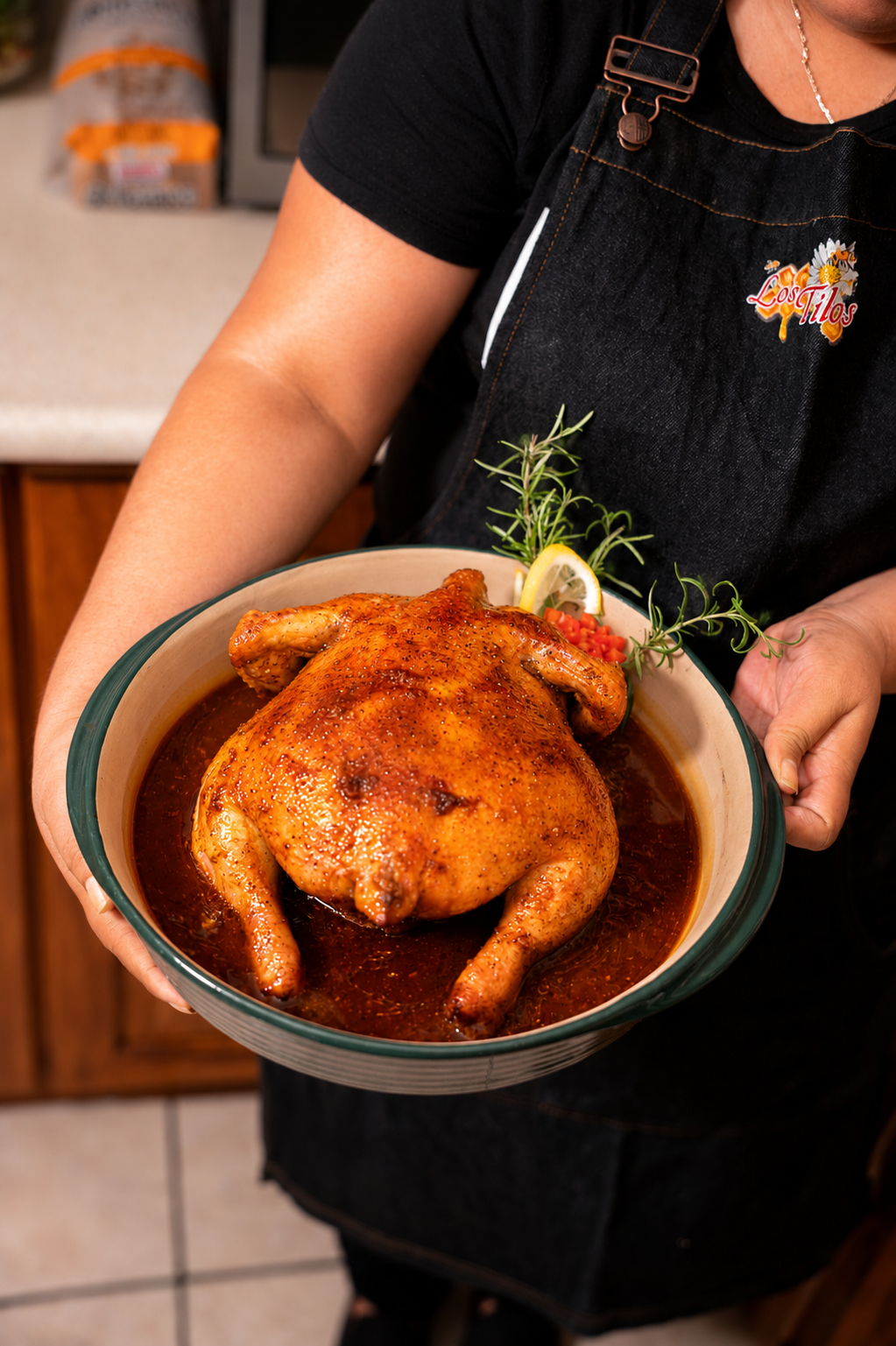 Persona sosteniendo un plato con pollo asado, decorado con ramitas de romero, una rodaja de limón y una pequeña porción de jitomates.