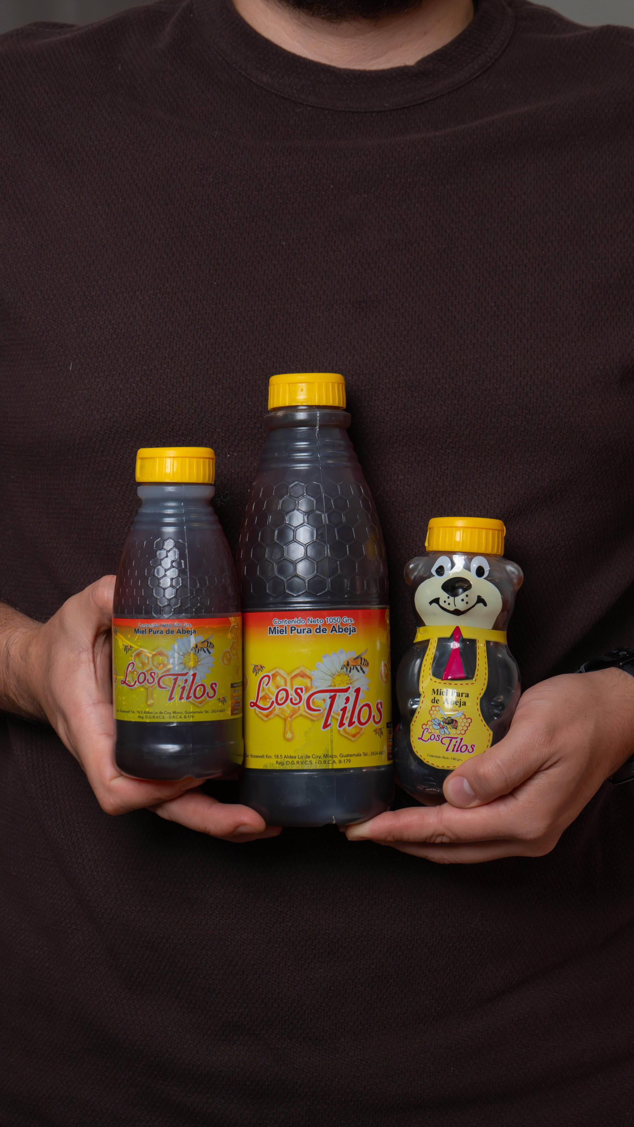 Persona sosteniendo tres botellas de miel pura de abeja 'Los Tilos', una con figura de oso, en fondo oscuro.