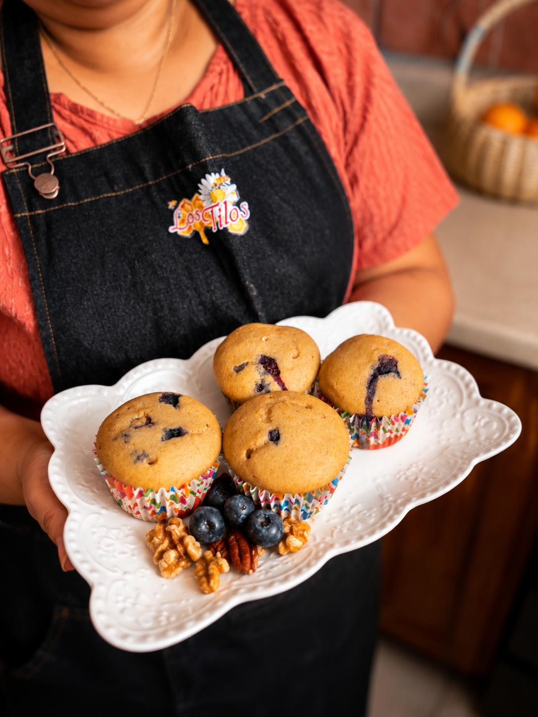 Persona sosteniendo un plato de muffins de arándano en una mesa, con un fondo de cesta con frutas. La persona lleva un delantal negro con un pin que dice "Los Filos" y una camiseta color coral.