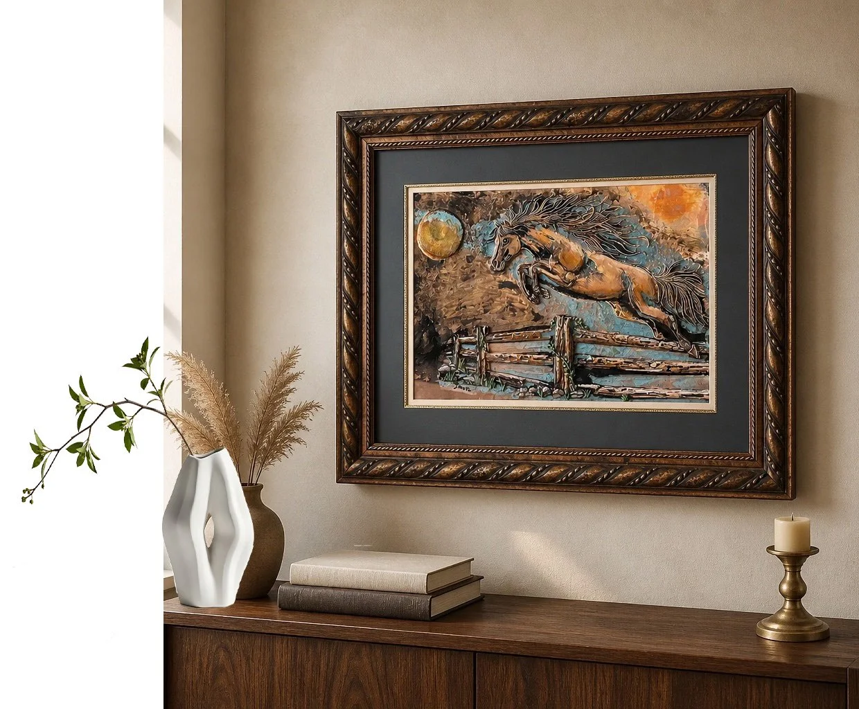 A framed painting of a horse jumping over a fence at sunset hangs on a beige wall, with a wooden sideboard below holding a white vase with greenery and pampas grass, two books, and a brass candle holder with a white candle.
