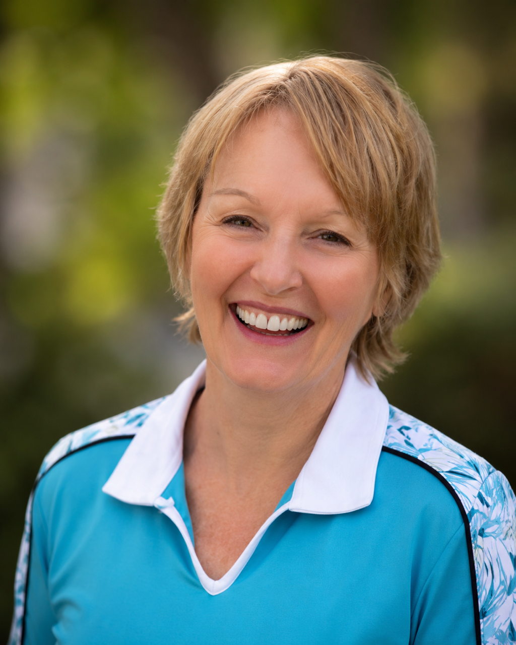 A smiling middle-aged woman with short blonde hair outdoors, wearing a blue sports shirt with a white collar and floral pattern on the shoulders.