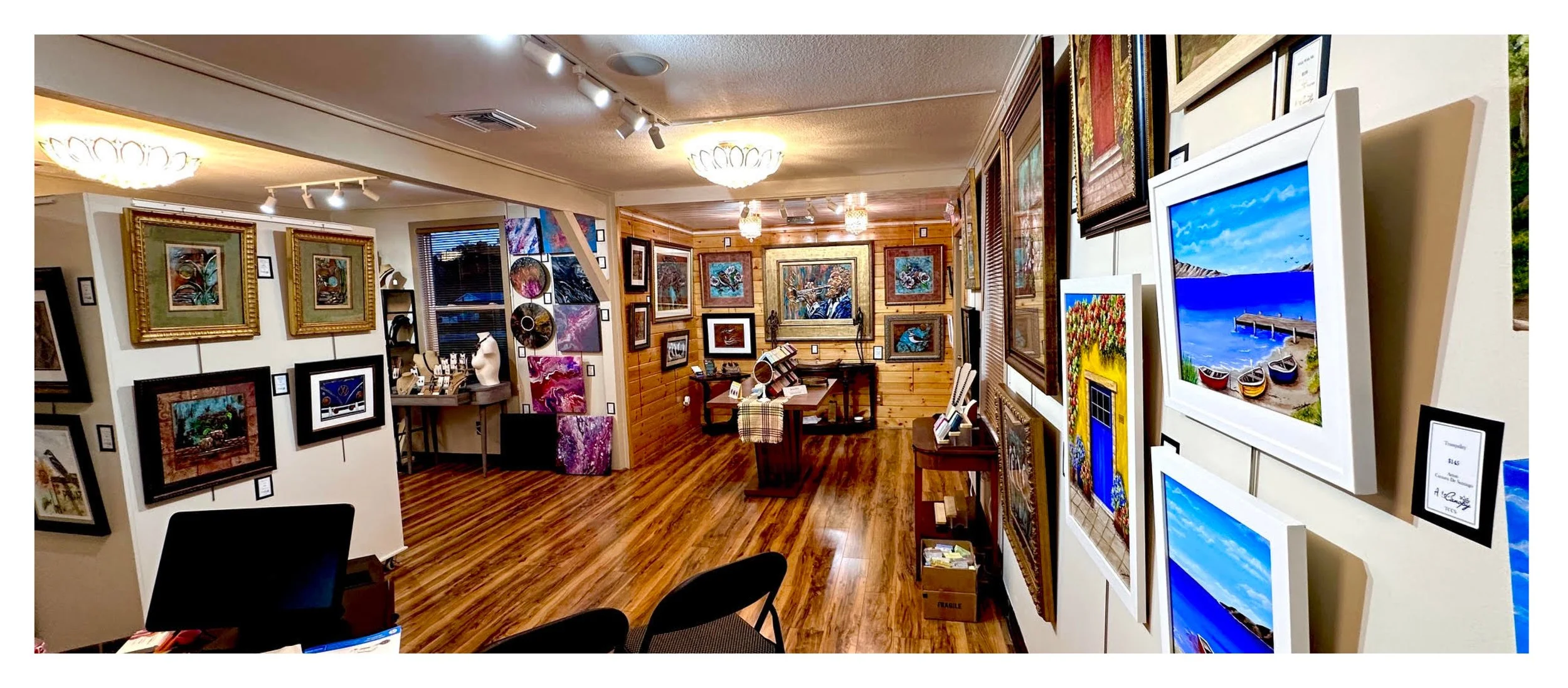 Interior of an art gallery with framed paintings and photographs displayed on white and wooden walls, including landscape artwork of boats on water and colorful abstract pieces, with a wooden floor and ceiling lights.