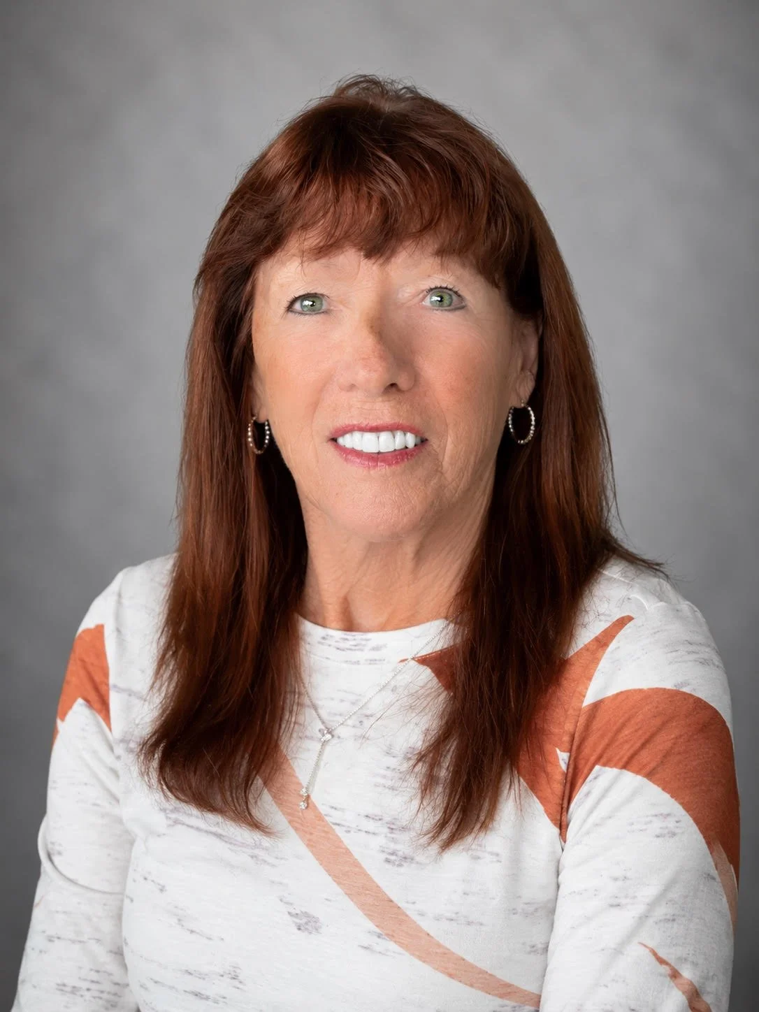 Portrait of a middle-aged woman with auburn hair, wearing hoop earrings, a white top with orange and gray patterns, smiling slightly against a plain gray background.