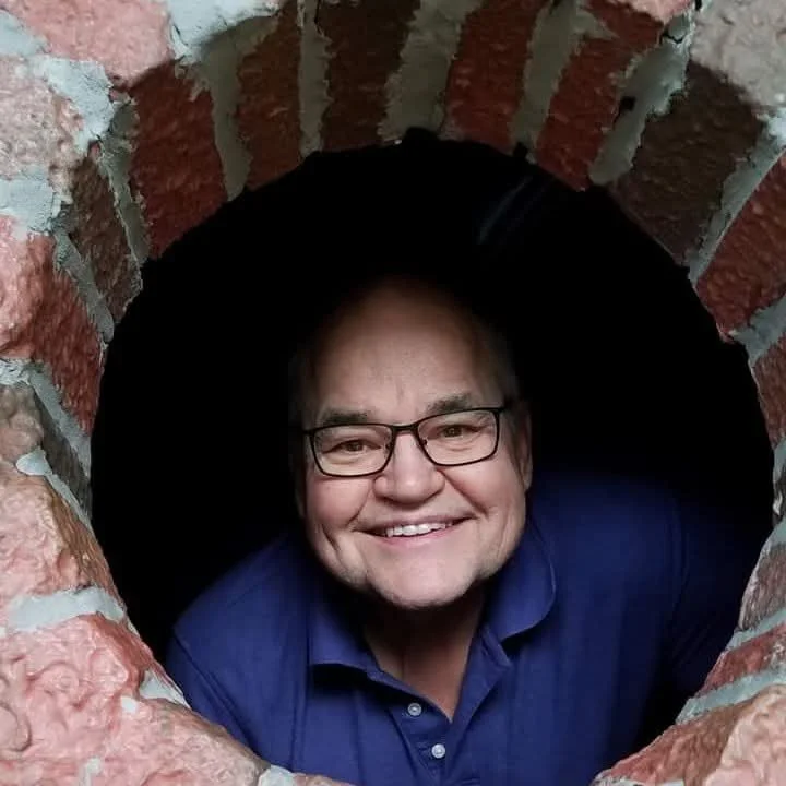 A man with glasses and a blue shirt smiling while looking through a round brick opening.