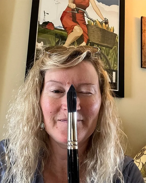 A woman with blonde curly hair and glasses on her head is holding a large paintbrush vertically in front of her face, covering her nose and mouth, in a room with framed artwork on the wall behind her.