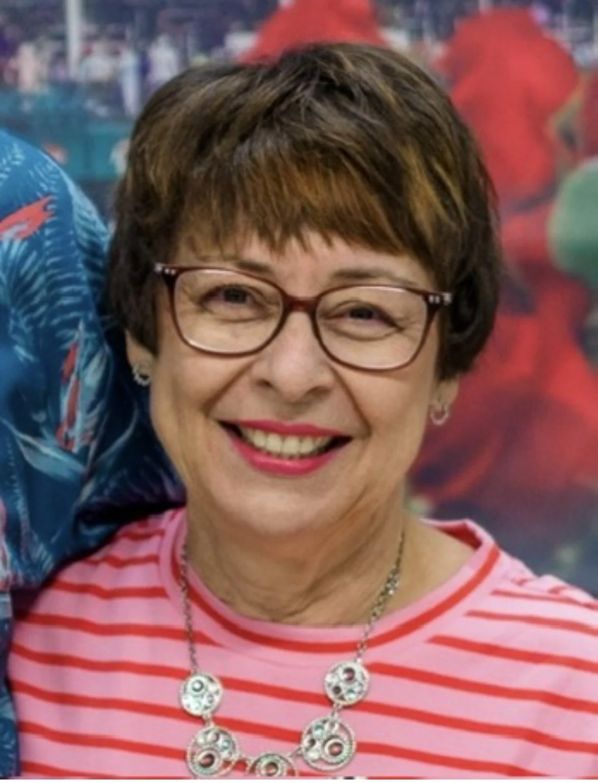 Close-up of woman with short brown hair, glasses, pink lipstick, wearing a red and white striped shirt and a silver necklace, smiling. Background is blurred and colorful.
