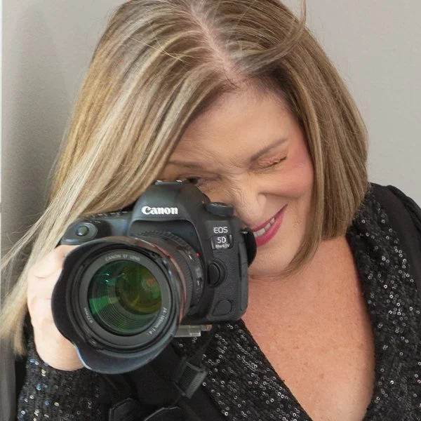 Woman with shoulder-length blonde hair, winking and smiling while holding a Canon EOS 5D Mark IV camera up to her face.