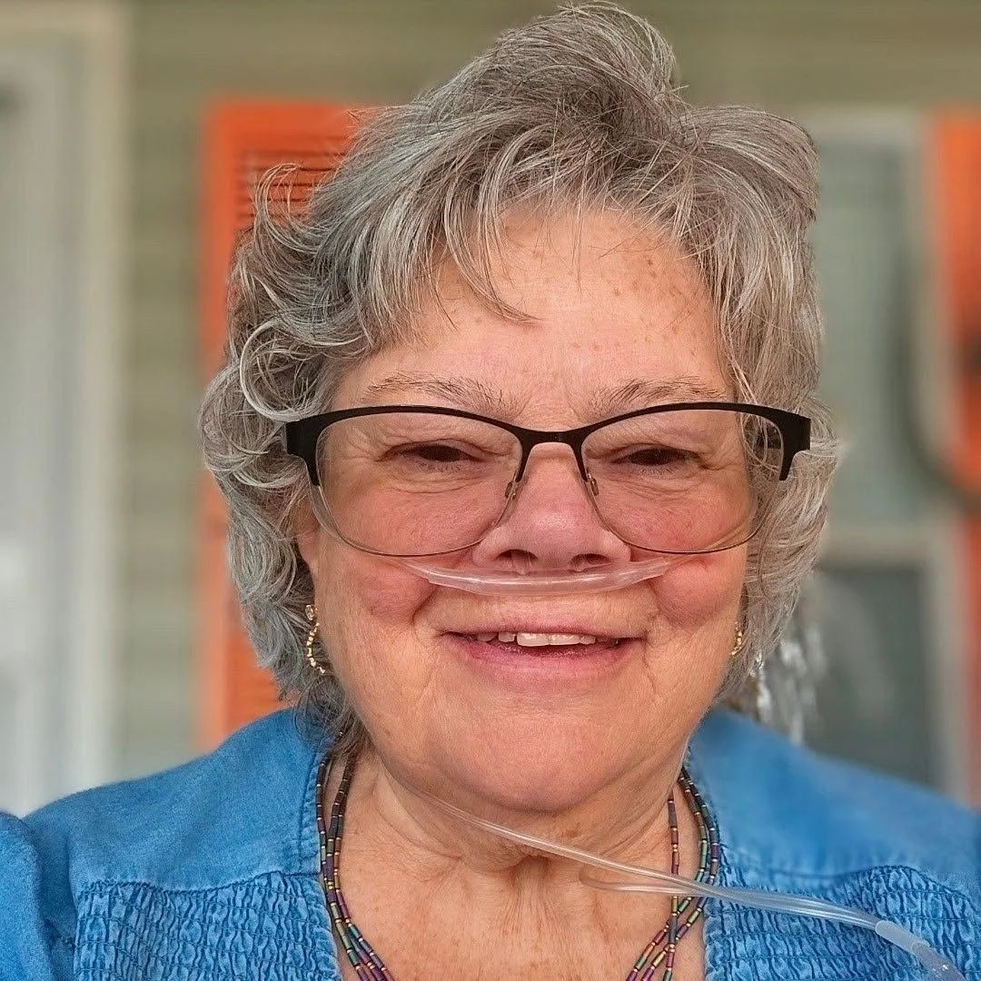 A smiling elderly woman with gray hair, glasses, and a nasal oxygen tube, wearing a blue top and beaded necklace, standing outdoors with blurred colorful background.