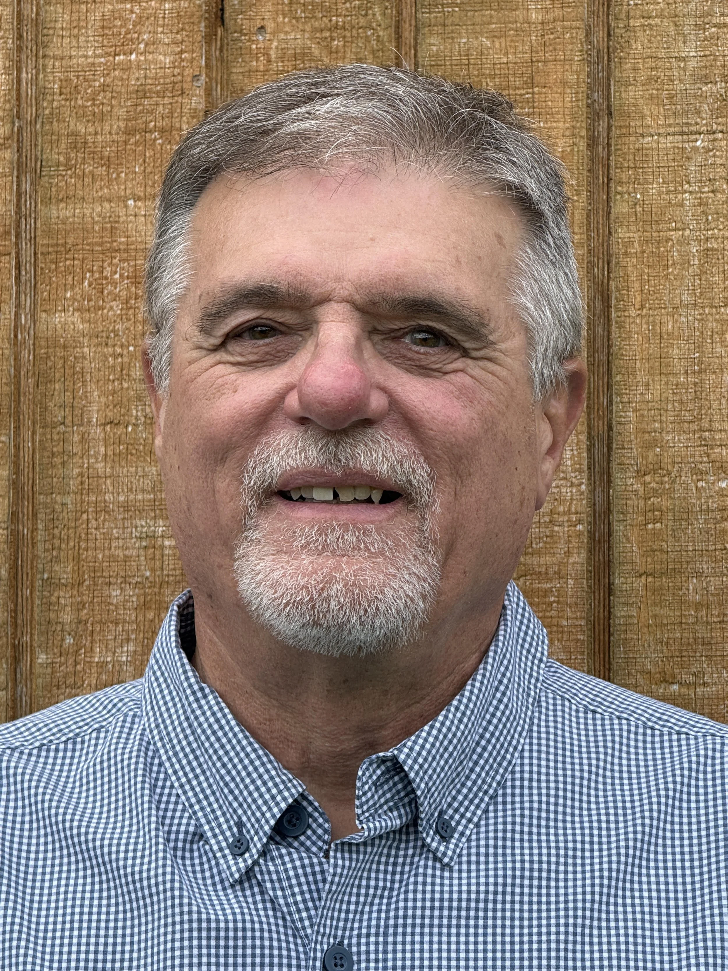 Close-up of a middle-aged man with gray hair and a gray beard, smiling, wearing a blue and white checkered shirt against a wooden background.