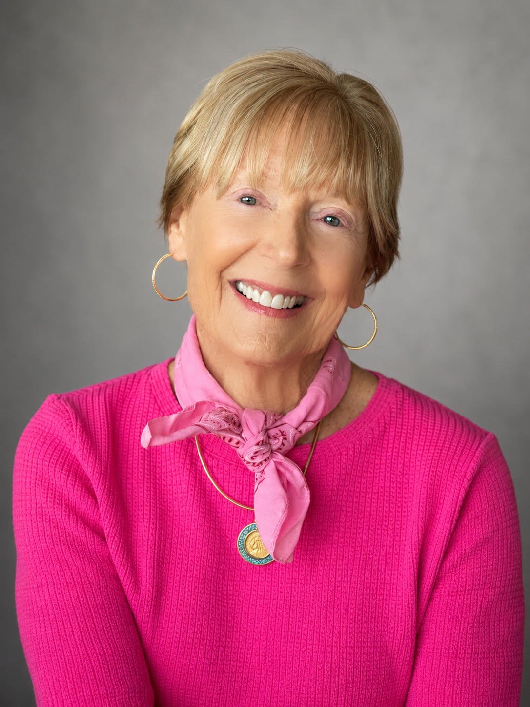 Portrait of an elderly woman smiling, wearing a pink sweater, a pink scarf with a bow, gold hoop earrings, and a gold necklace with a pendant, against a neutral gray background.