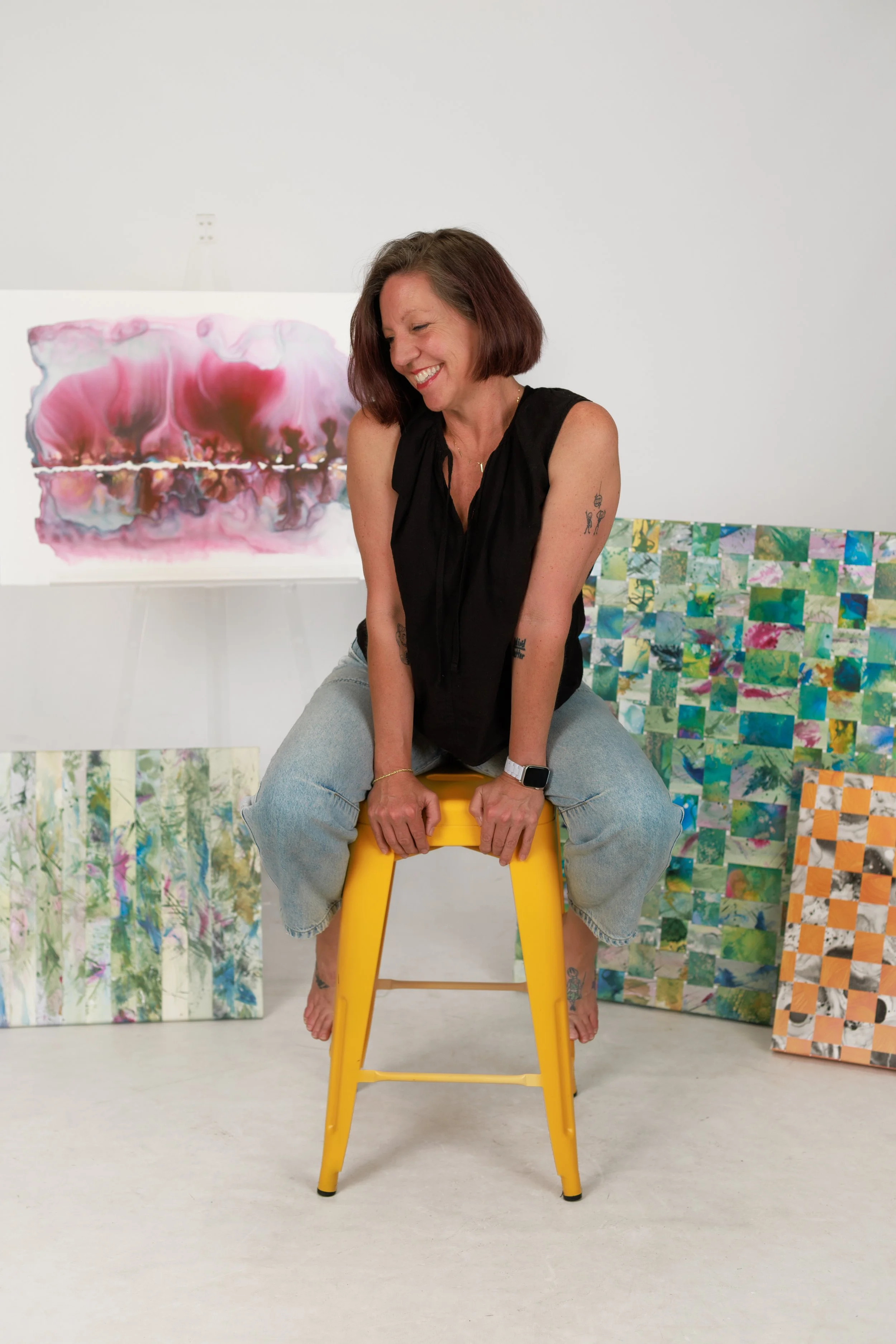 Woman with short brown hair sitting on a yellow step stool in front of abstract and patterned paintings in an art studio, smiling with eyes closed, wearing a black sleeveless top and light blue jeans.