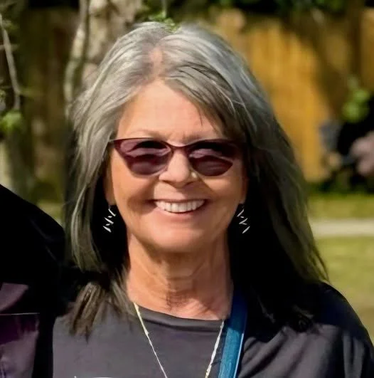 A smiling woman with gray hair wearing sunglasses, earrings, and a black top outdoors.