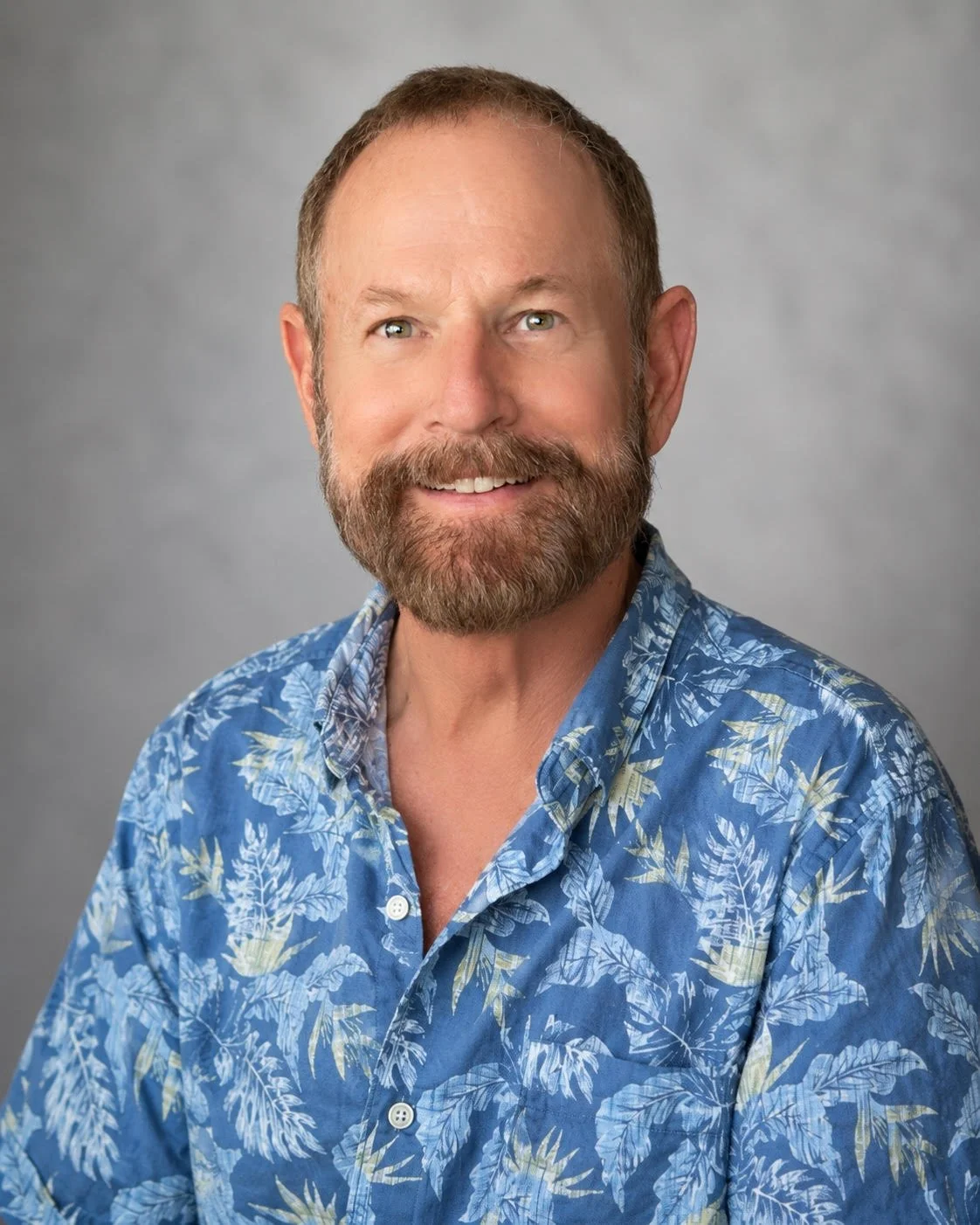 Headshot of a middle-aged man with a beard and short hair, wearing a blue floral shirt against a gray background.