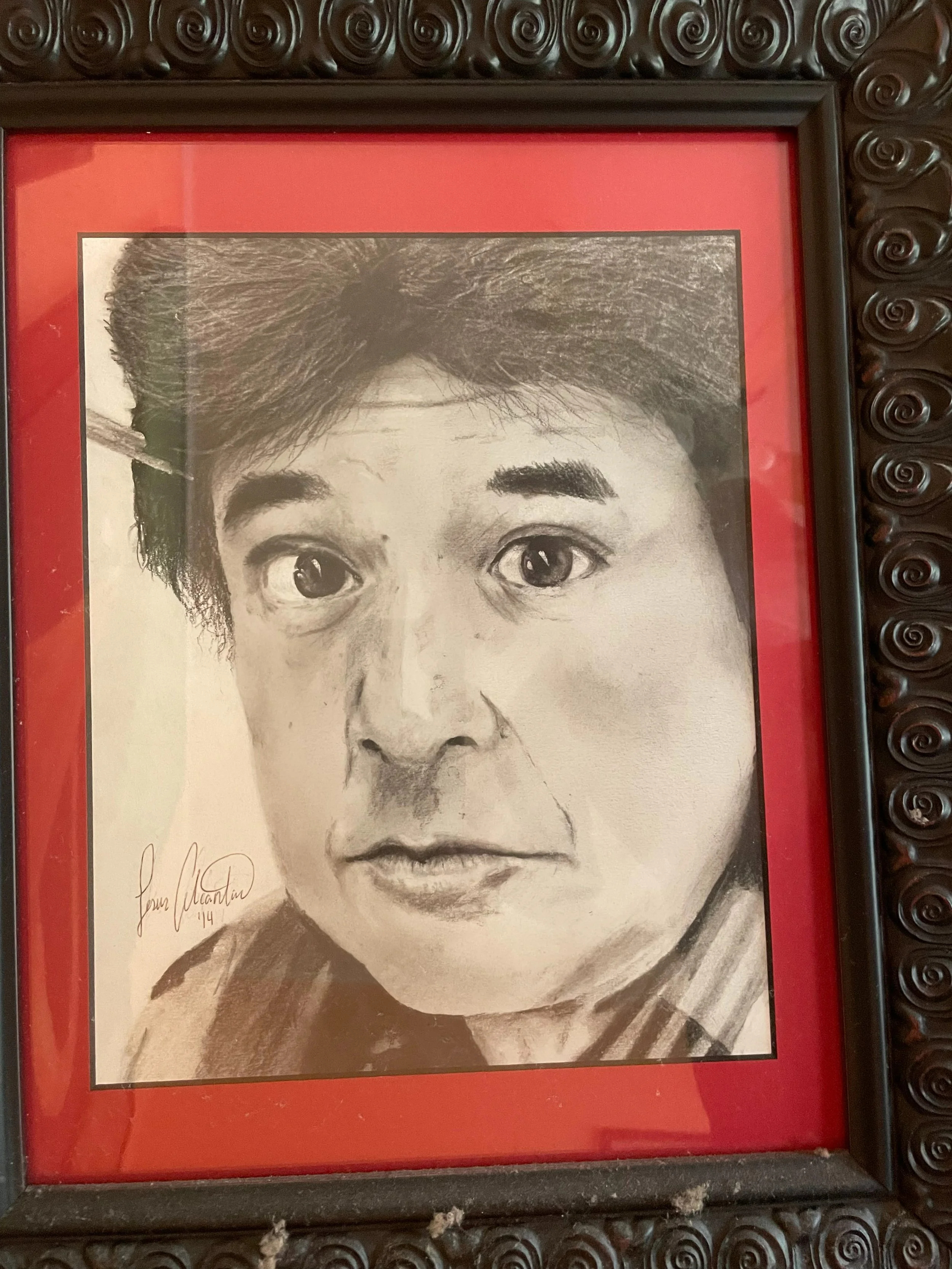 A black-and-white portrait of a person with short hair, expressive eyebrows, and serious expression, framed with a red mat and decorative black frame.