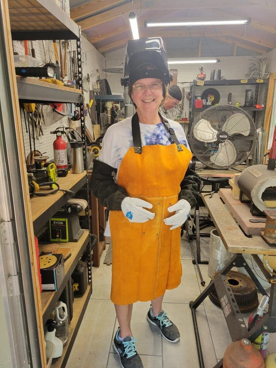 An older woman in a workshop, wearing a welding helmet pushed back on her head, safety gloves, an orange apron, a white t-shirt, black long sleeves, sneakers, and smiling at the camera.