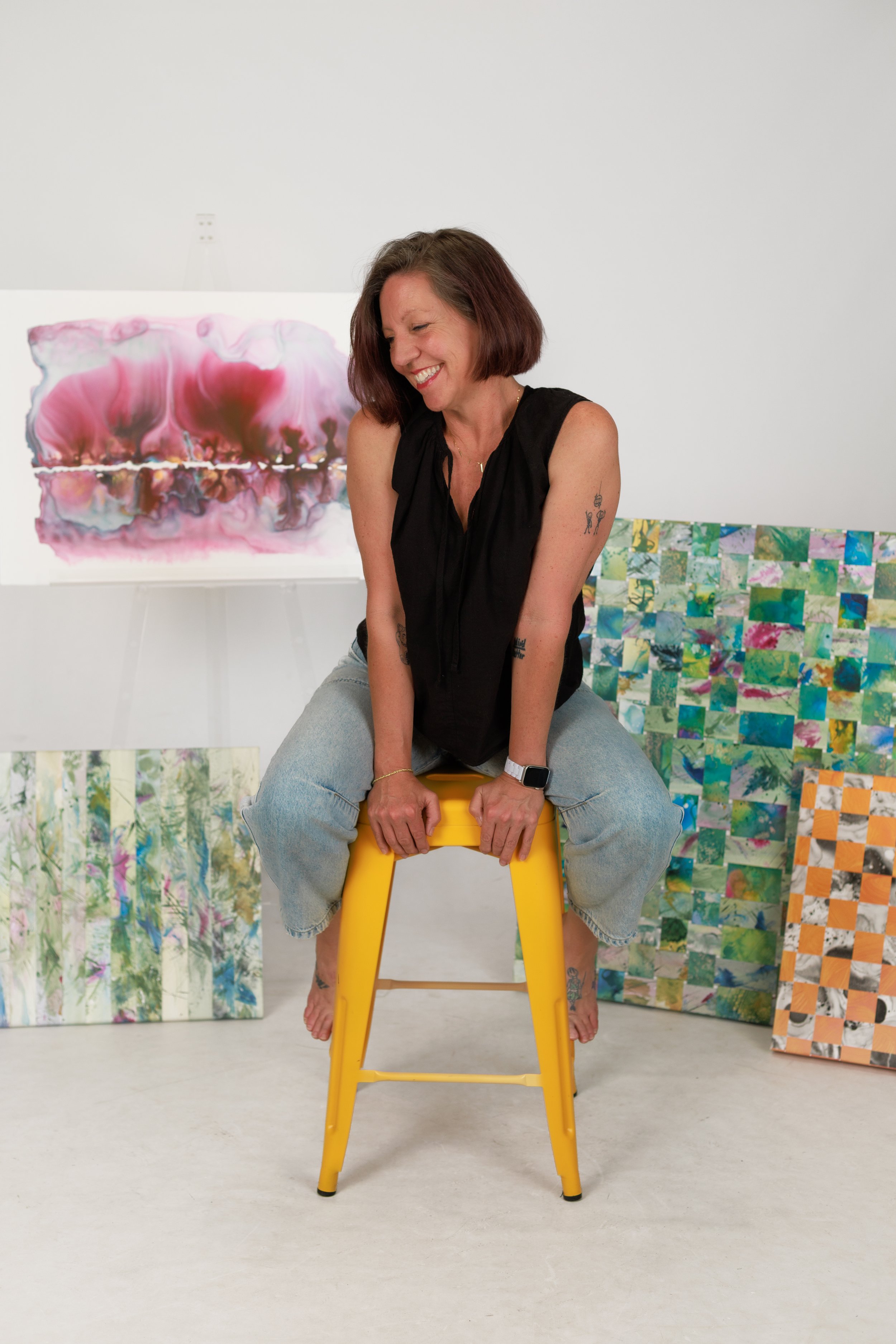A smiling woman with shoulder-length brown hair, wearing a black sleeveless top and light blue jeans, sitting on a yellow chair in front of abstract and patterned artworks displayed on the wall behind her.