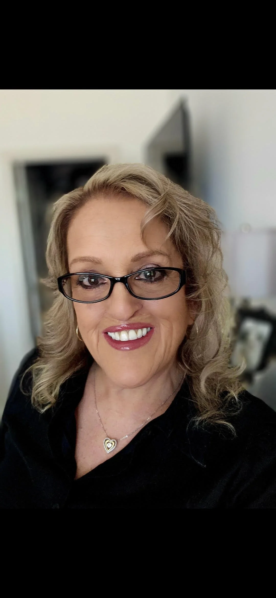 A close-up of a middle-aged woman with blonde hair, glasses, and a bright smile, wearing a black top and a heart-shaped necklace, in a well-lit indoor setting.