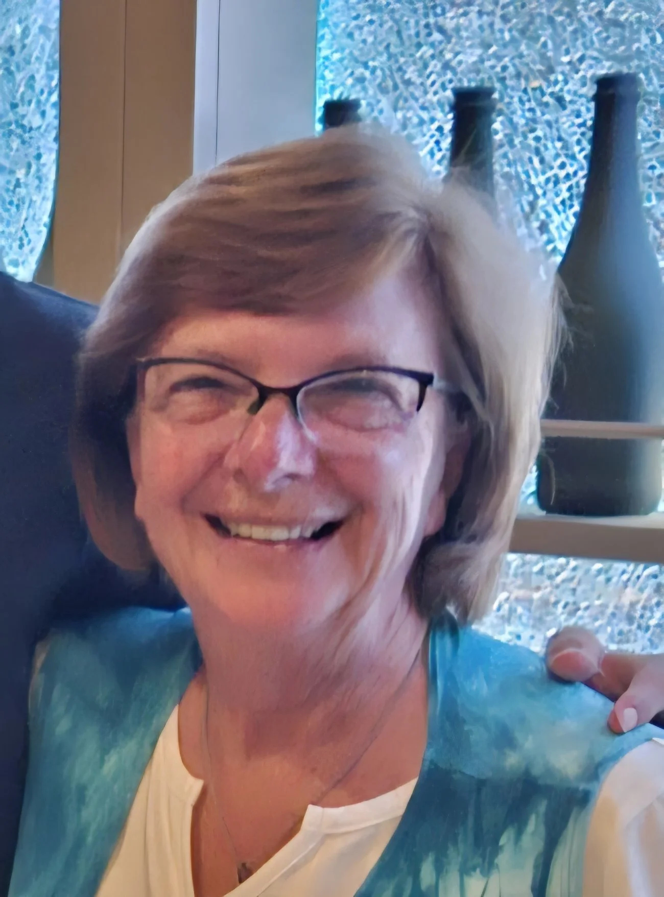 A smiling older woman with glasses and short blonde hair, wearing a light blue and white top, sitting outdoors with a blue textured wall and Vases in the background.