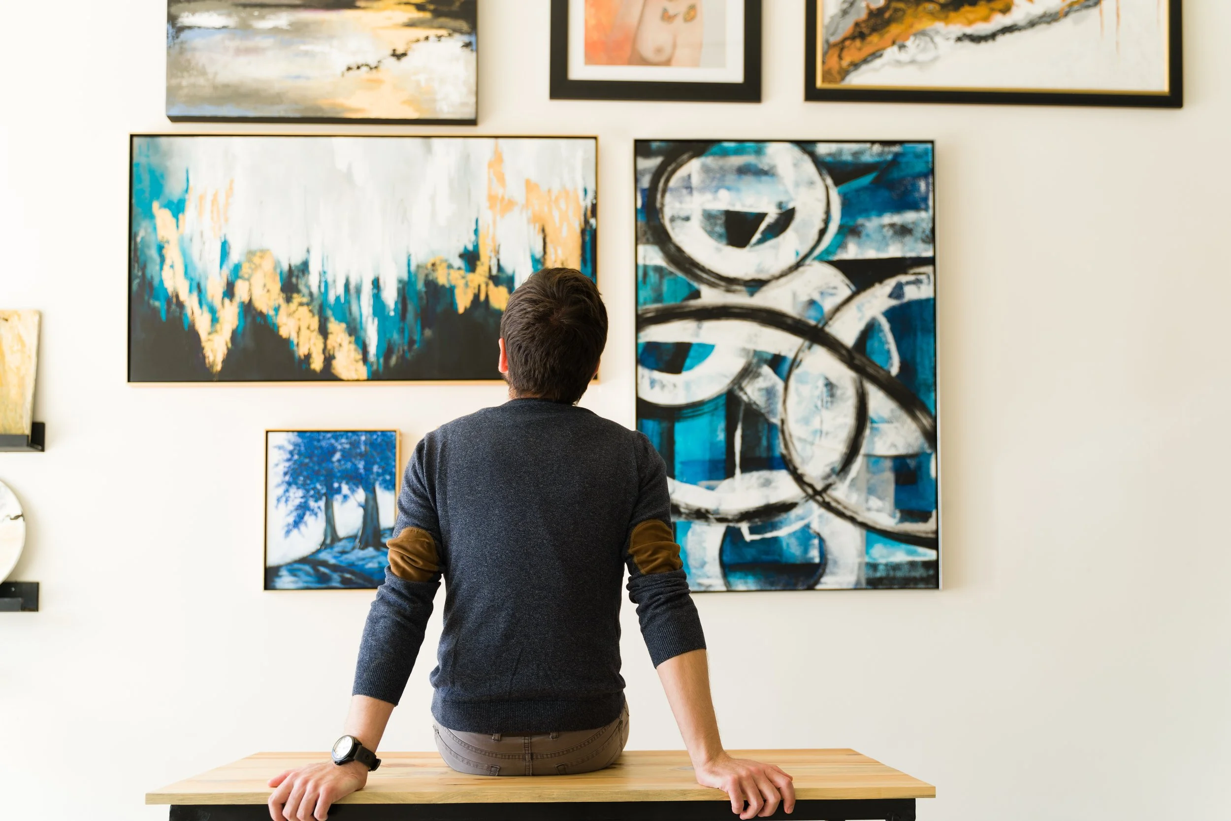 A man with brown hair, wearing a dark sweater, is standing in front of a white wall with various abstract paintings. His back is turned to the camera and he is leaning on a wooden table, looking at the artwork.