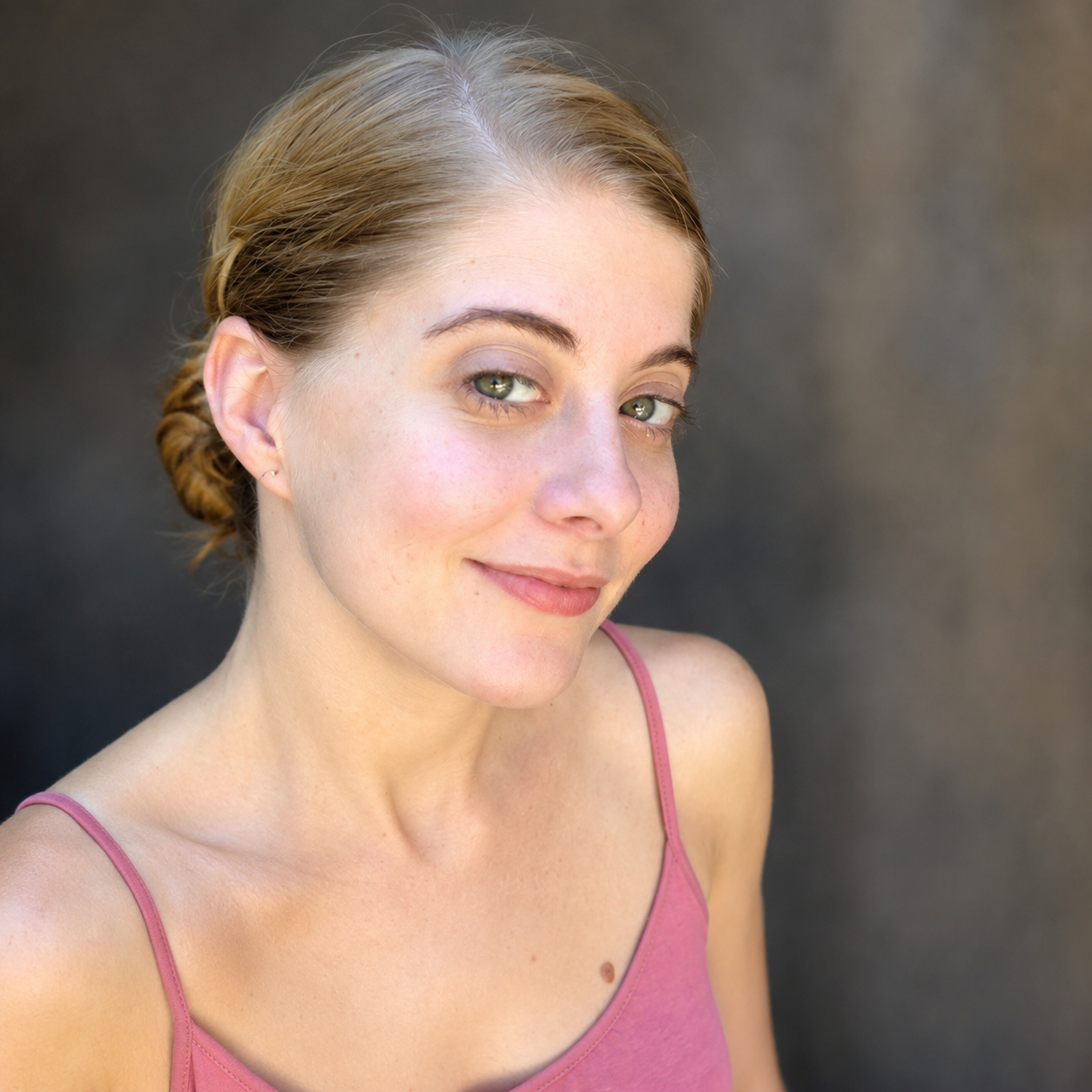 A close-up of a young woman with light brown hair styled in a low bun, wearing a pink tank top, smiling and looking at the camera, against a dark, blurred background.