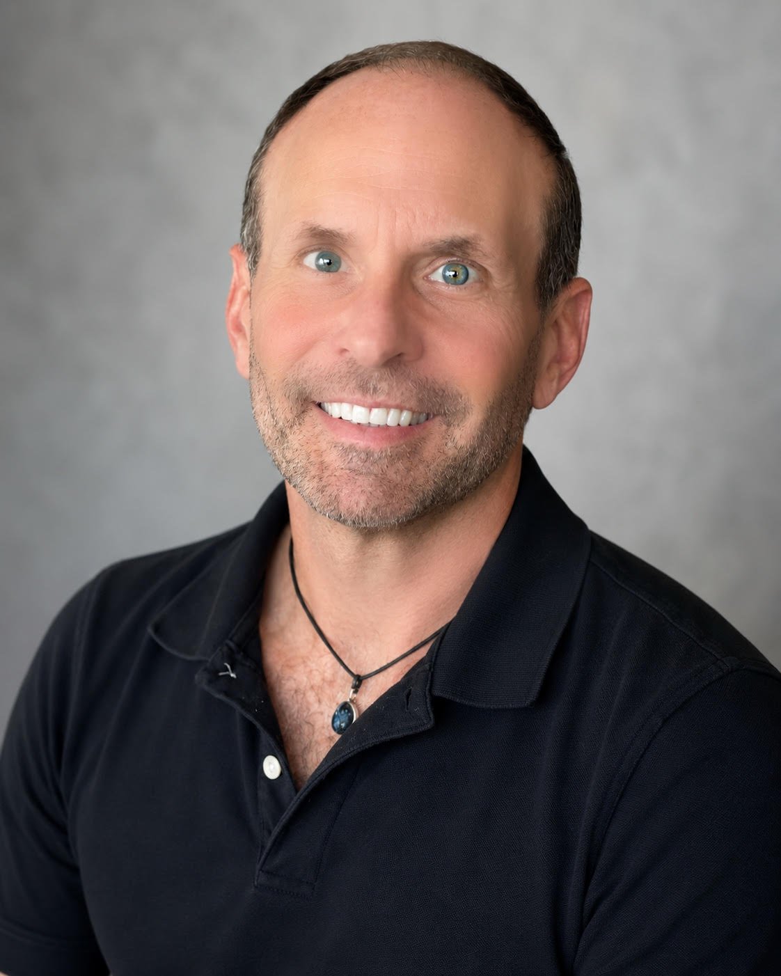 Headshot of a middle-aged man with short brown hair, blue eyes, and a beard, smiling and wearing a black collared shirt with a necklace, against a gray background.