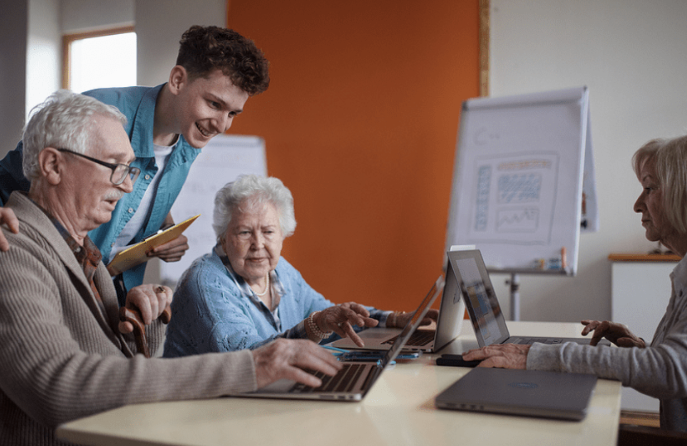 Young man and elderly adults working on laptops together in a classroom or meeting room.