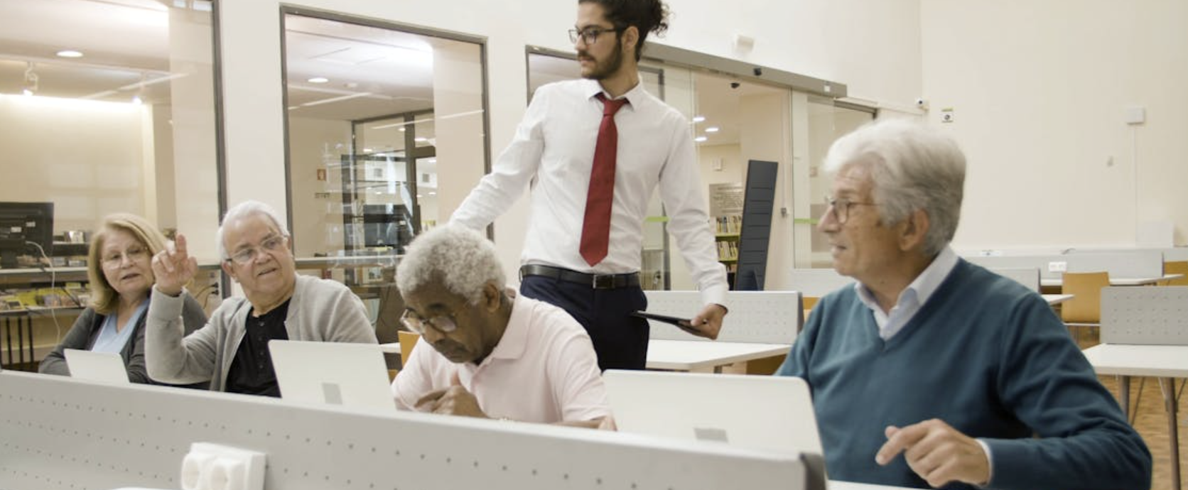 A young man with curly hair, glasses, wearing a white dress shirt and red tie, standing and talking to a group of elderly people sitting at tables in a library or classroom setting.