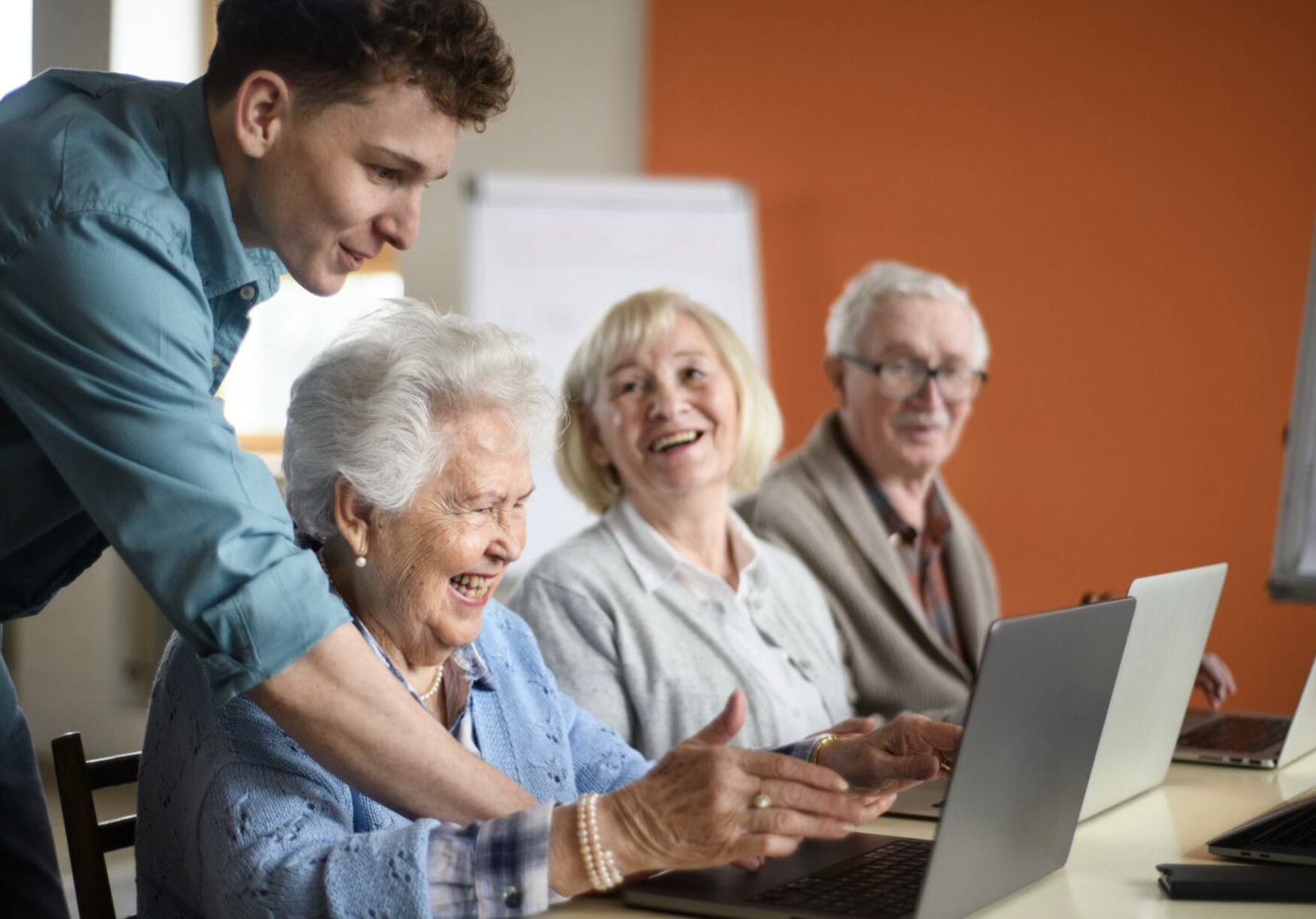 Young man helping elderly woman with laptop in a group setting, smiling and interacting.