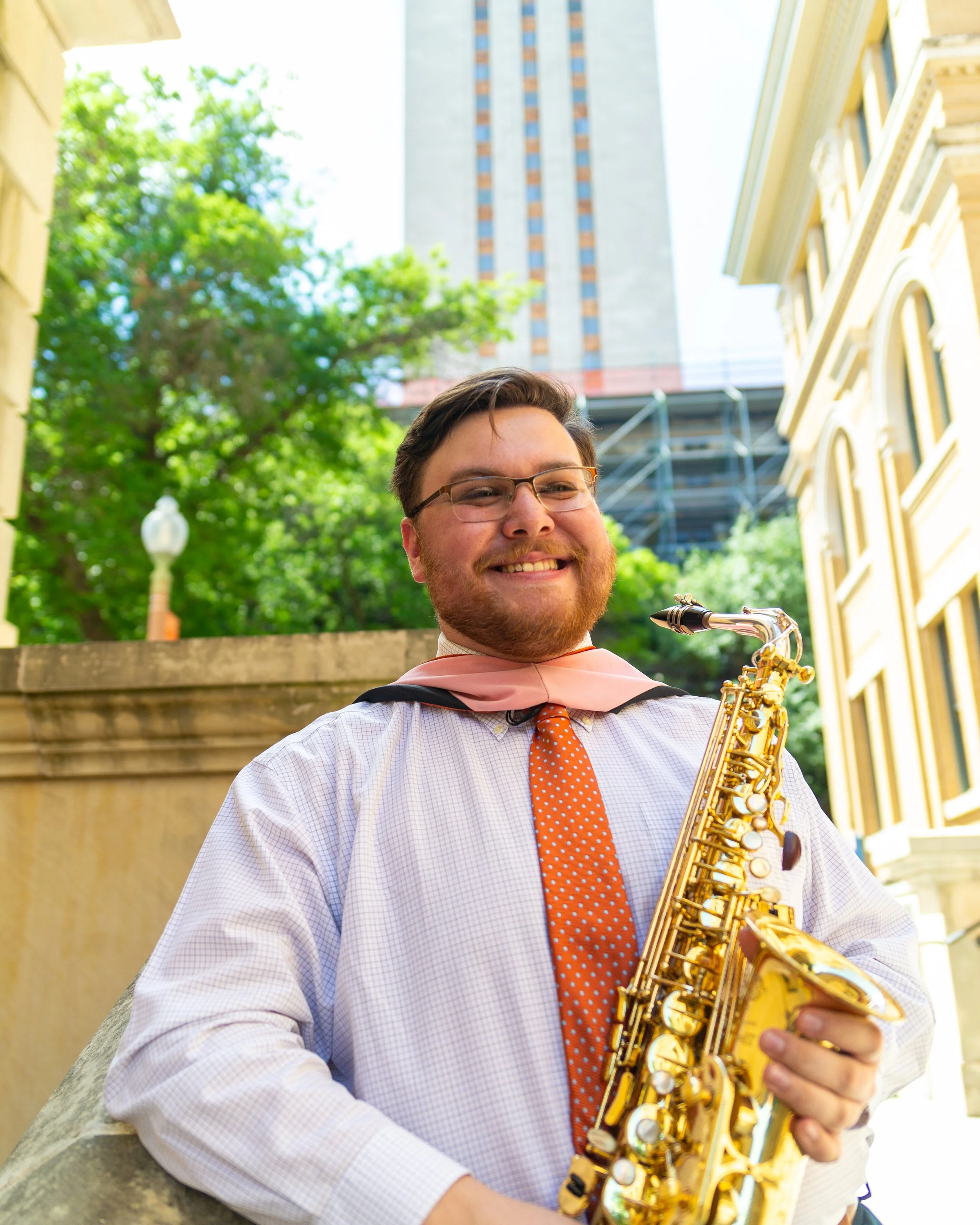 Male saxophonist smiling outdoors with green trees and buildings in the background.