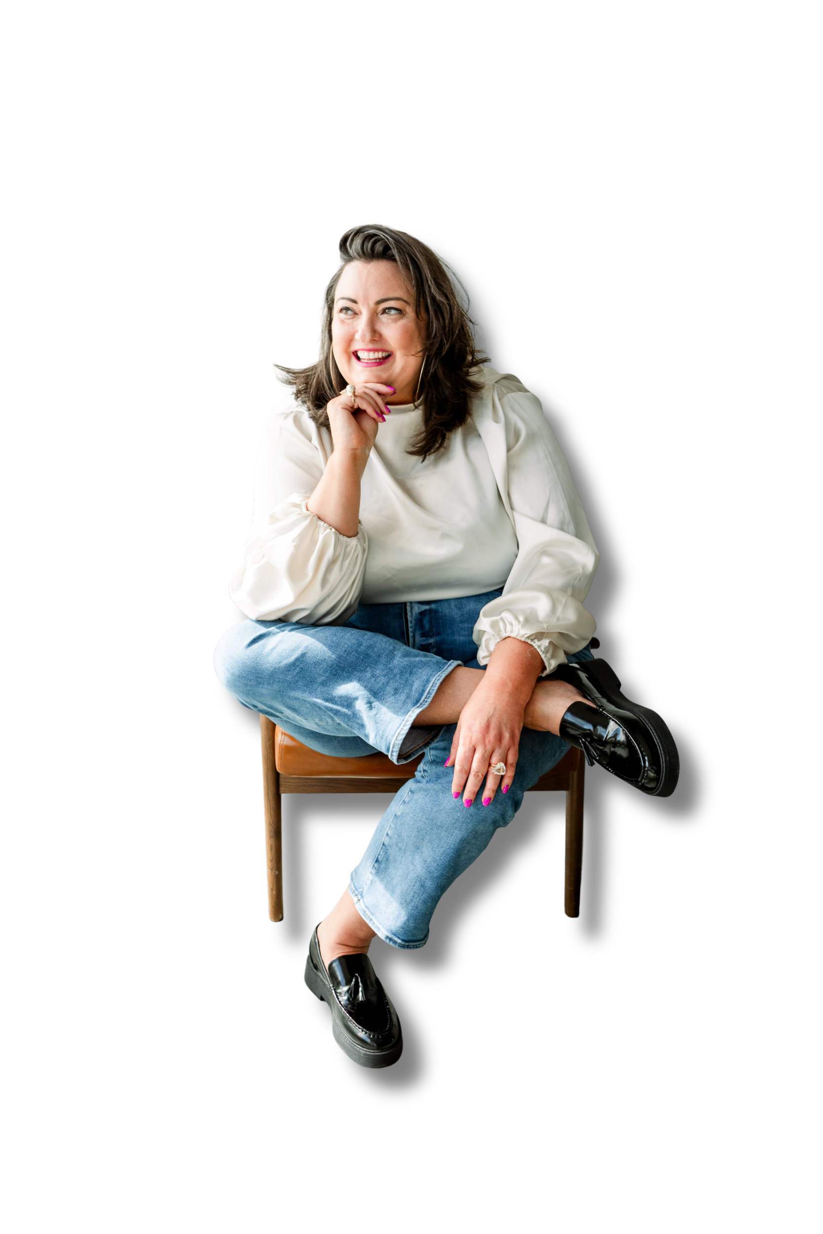 Summer Carrick, wearing a white blouse and blue jeans, sitting on a wooden chair. She has a cheerful expression, smiling and looking to her right. She is accessorized with rings and has pink nail polish, and is wearing black shiny loafers.