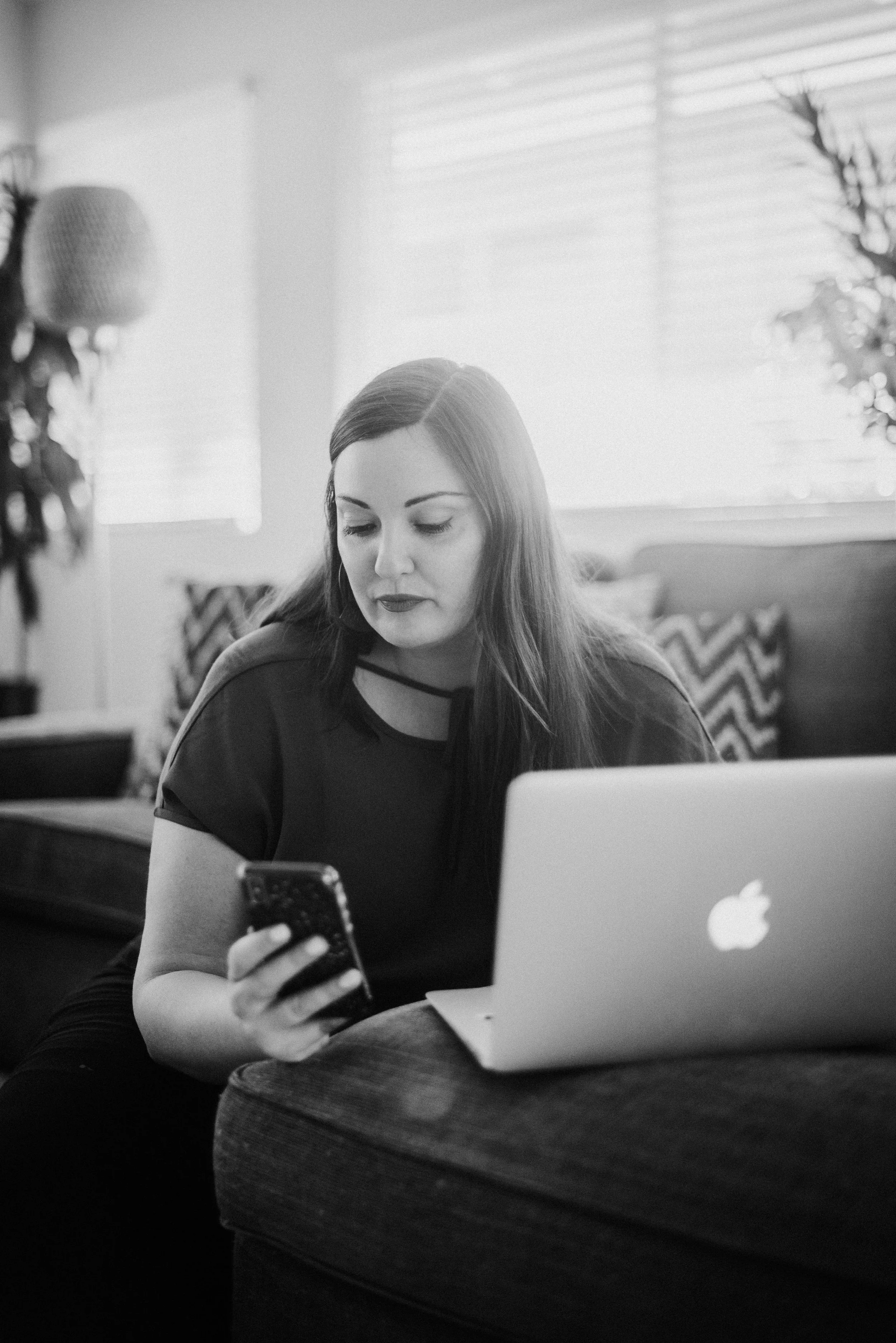 Summer Carrick with long hair looking at her smartphone to check on client social media accounts while sitting in front of an open laptop in a bright room in Kona.
