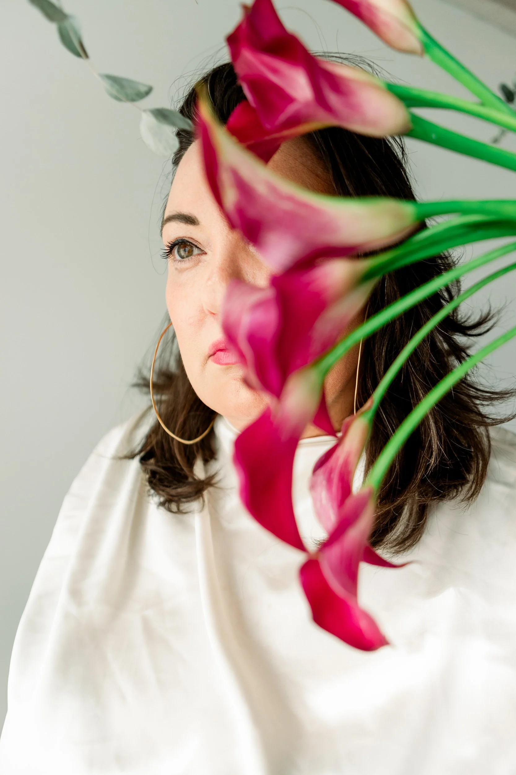 Summer Carrick on the photo set with big hoop earrings, wearing a white blouse, partially obscured by a bouquet of pink calla lilies and eucalyptus leaves, against a plain light background.
