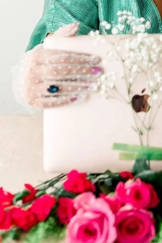 A person in a green top holds a white box with flowers on top, surrounded by pink and red flowers on a table.