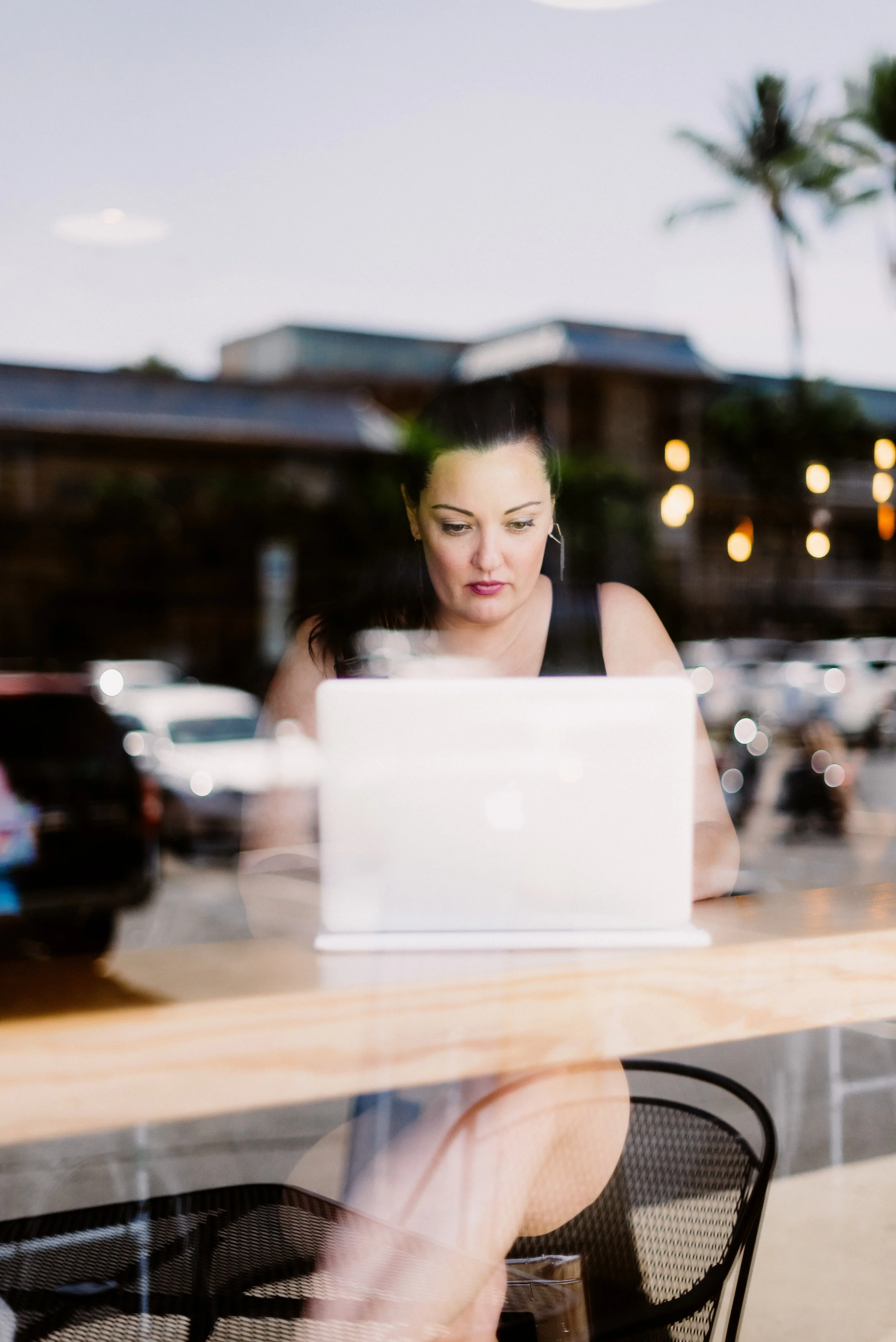 Summer Carrick, Marketer, at a Big Island cafe, sitting at a wooden table looking at a laptop, with blurred cars and palm trees in the background.