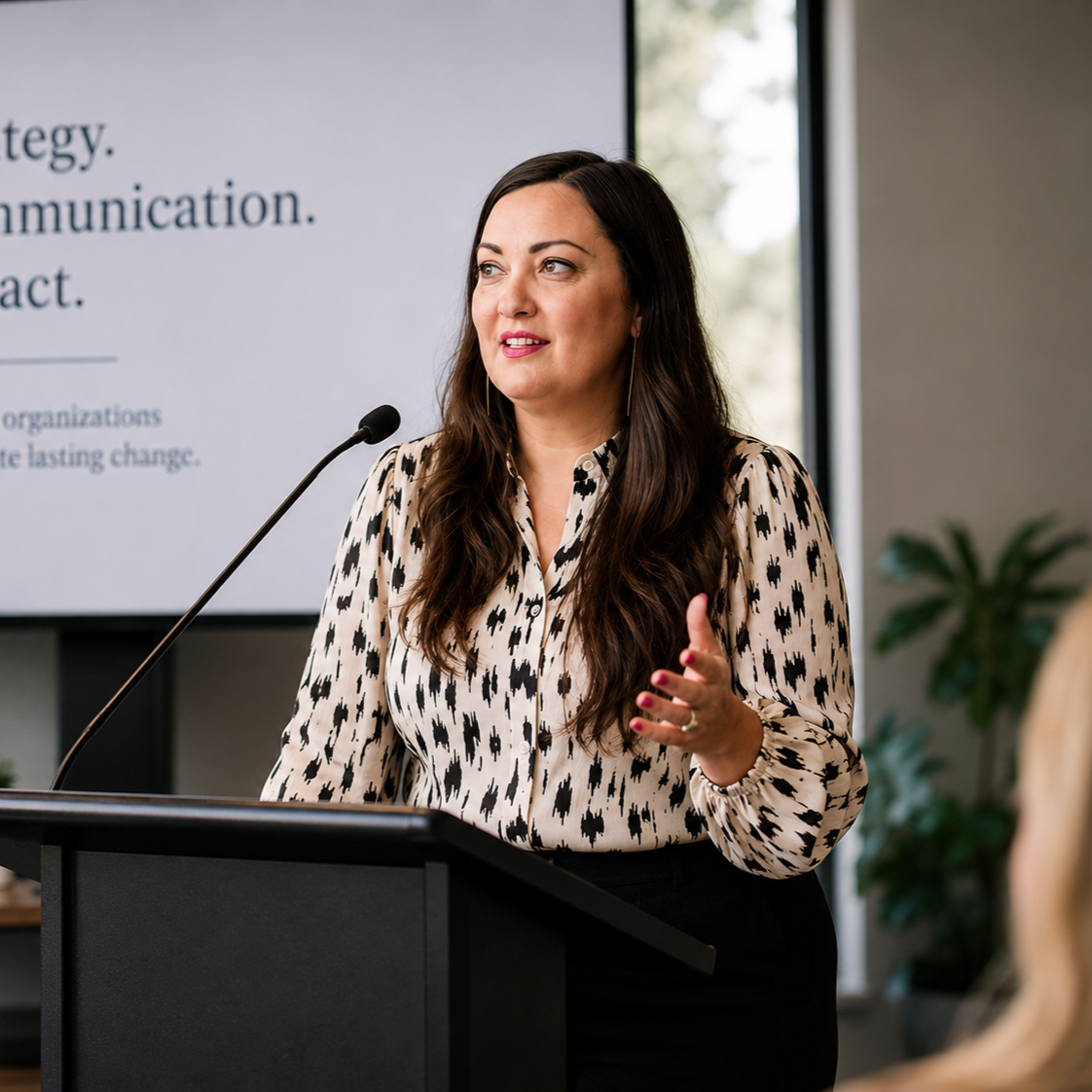 A woman with long dark hair giving a speech at a podium with a microphone, in a professional setting with a presentation screen in the background.