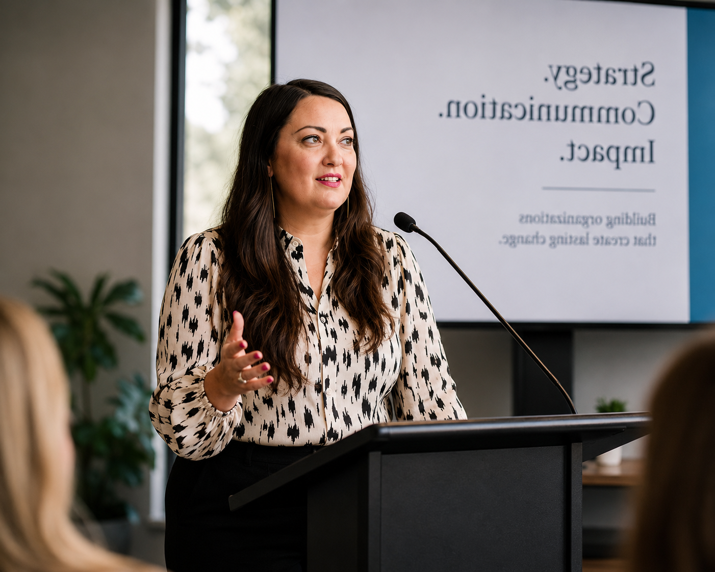 A woman with long dark hair and wearing a black and white patterned blouse speaking at a podium during a presentation, with a large screen behind her displaying a slide titled 'Strategy. Communication. Impact.' and a plant in the background.